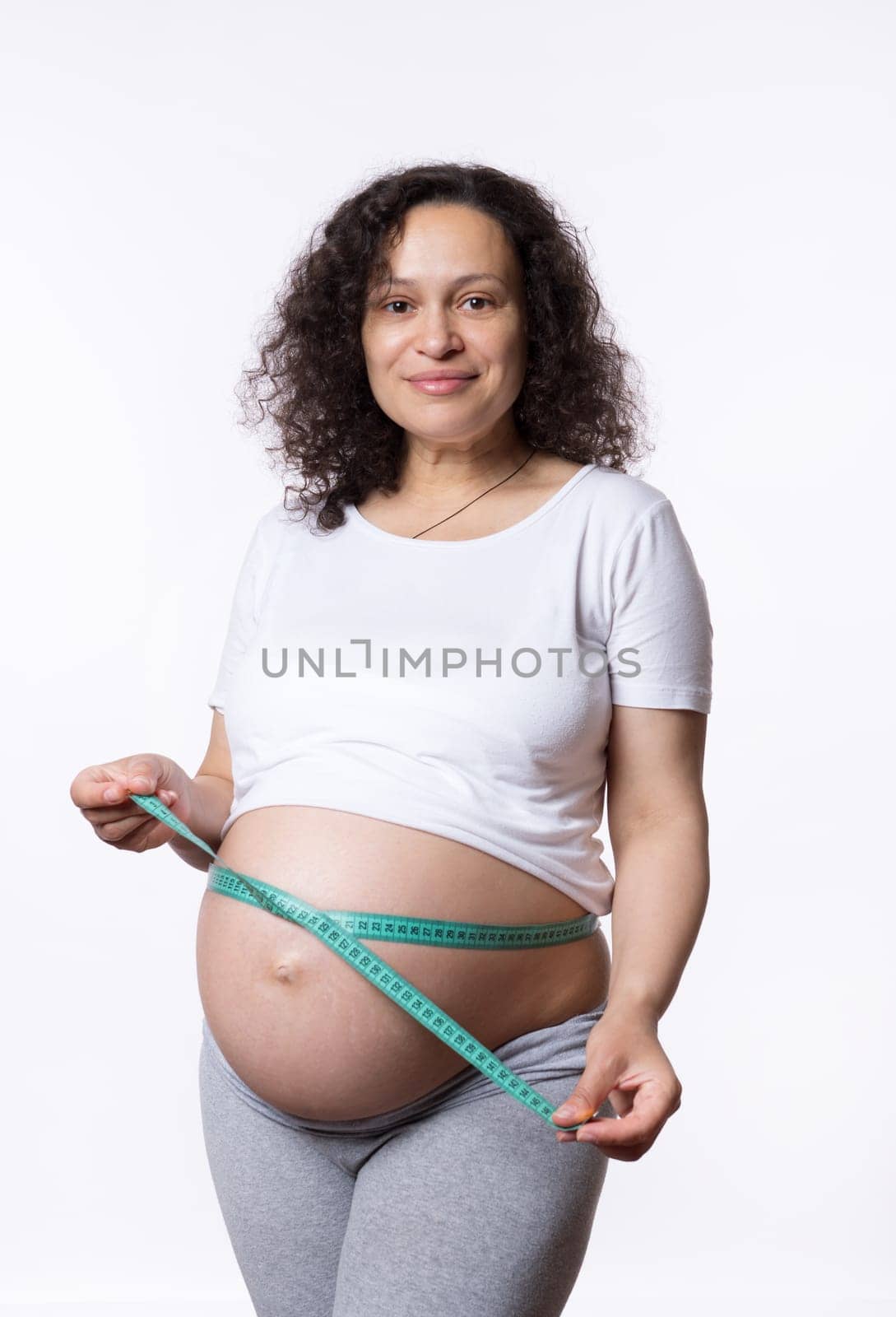 Beautiful gravid pregnant woman, expectant mother measuring her big belly in third trimester with tape, isolated white studio background. Pregnancy. Maternity. Female health and gynecology concept