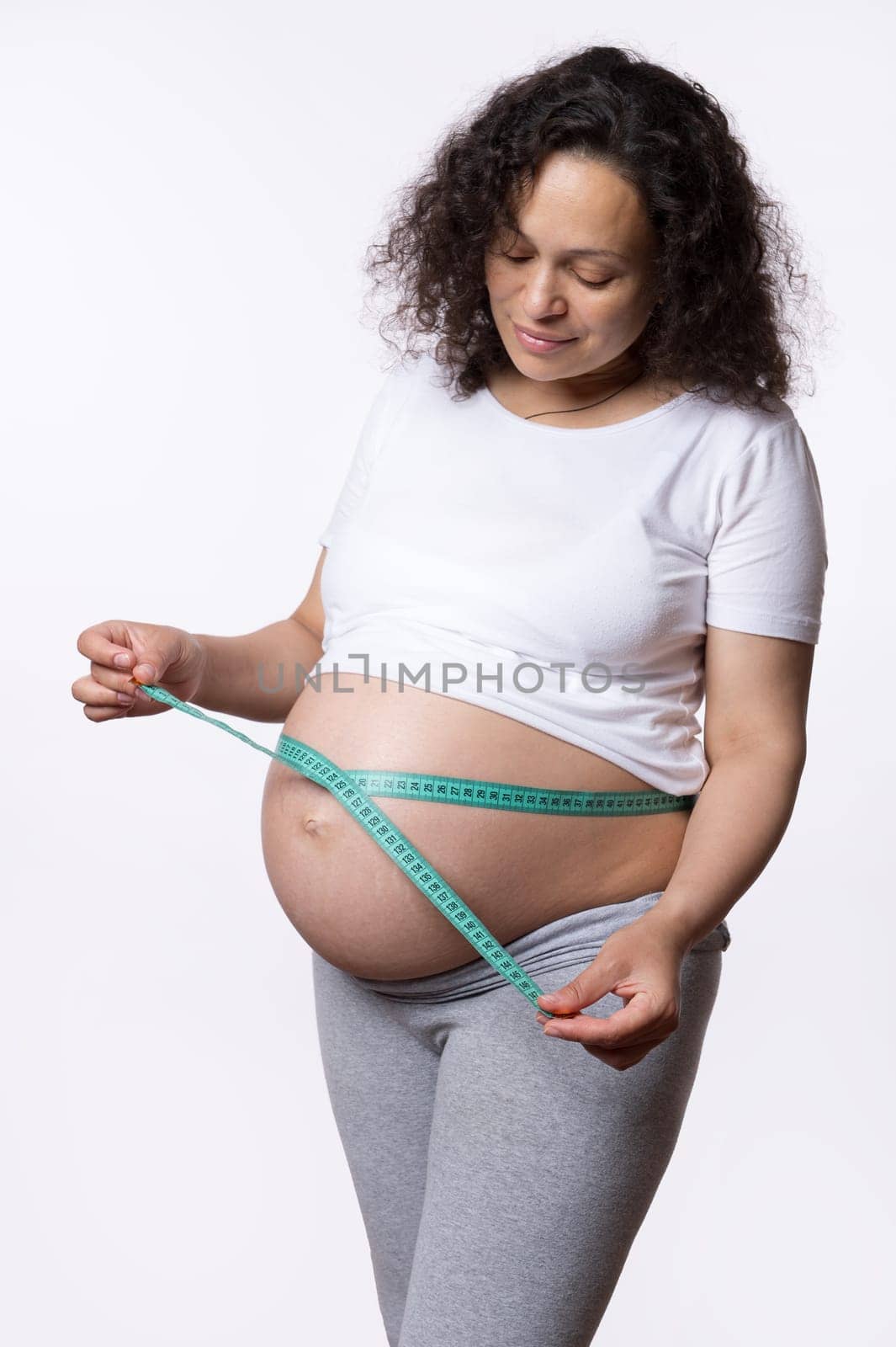 Multi-ethnic gravid woman measures her stomach with measuring tape over white background. Pregnant belly measurement. Baby health monitoring concept. Pregnancy. Maternity. Women's health and body care