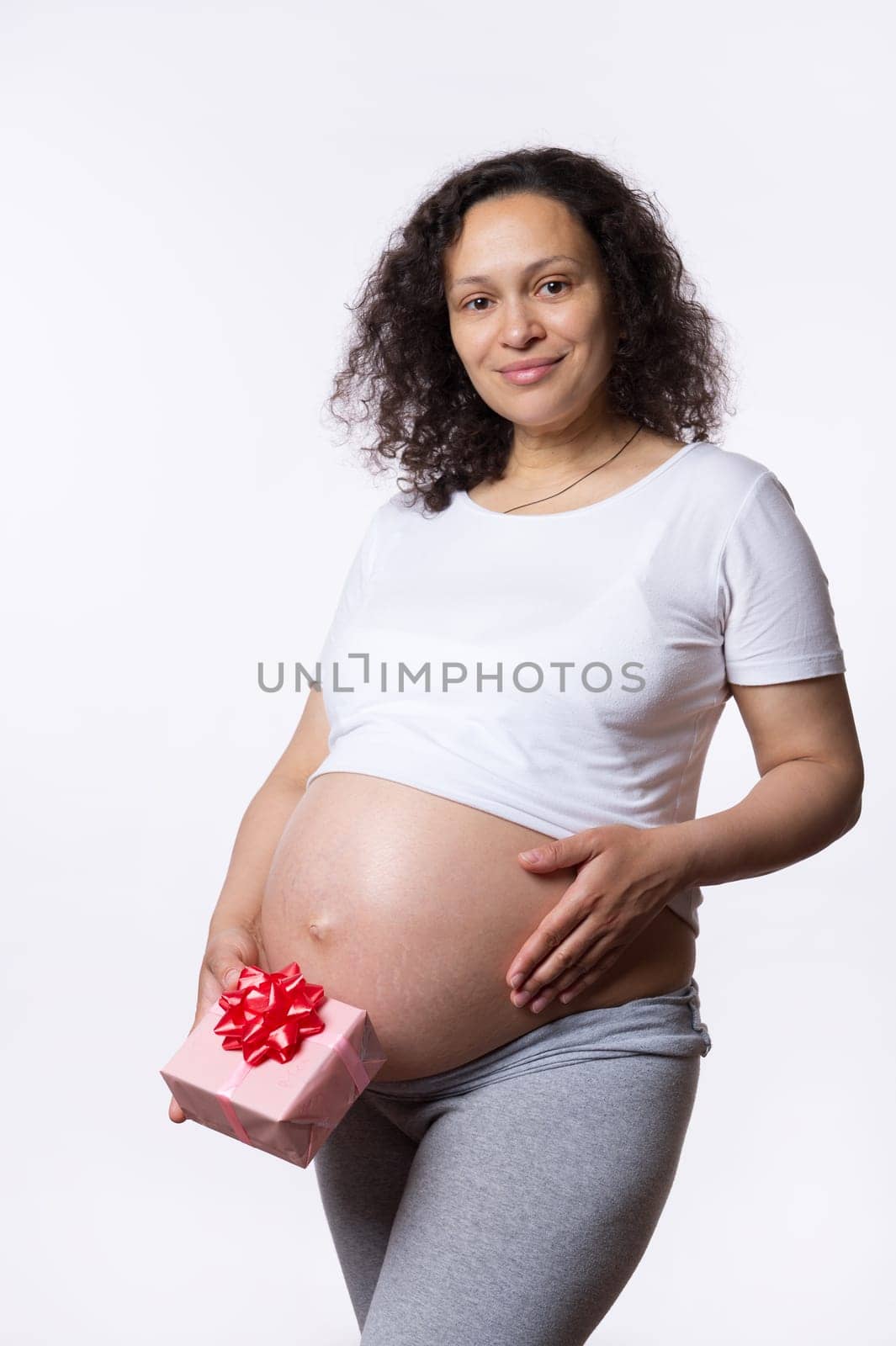 Attractive positive multi ethnic pregnant woman holding a gift box, touching and stroking her big belly in late pregnancy, smiling looking at camera, isolated over white studio background. Maternity