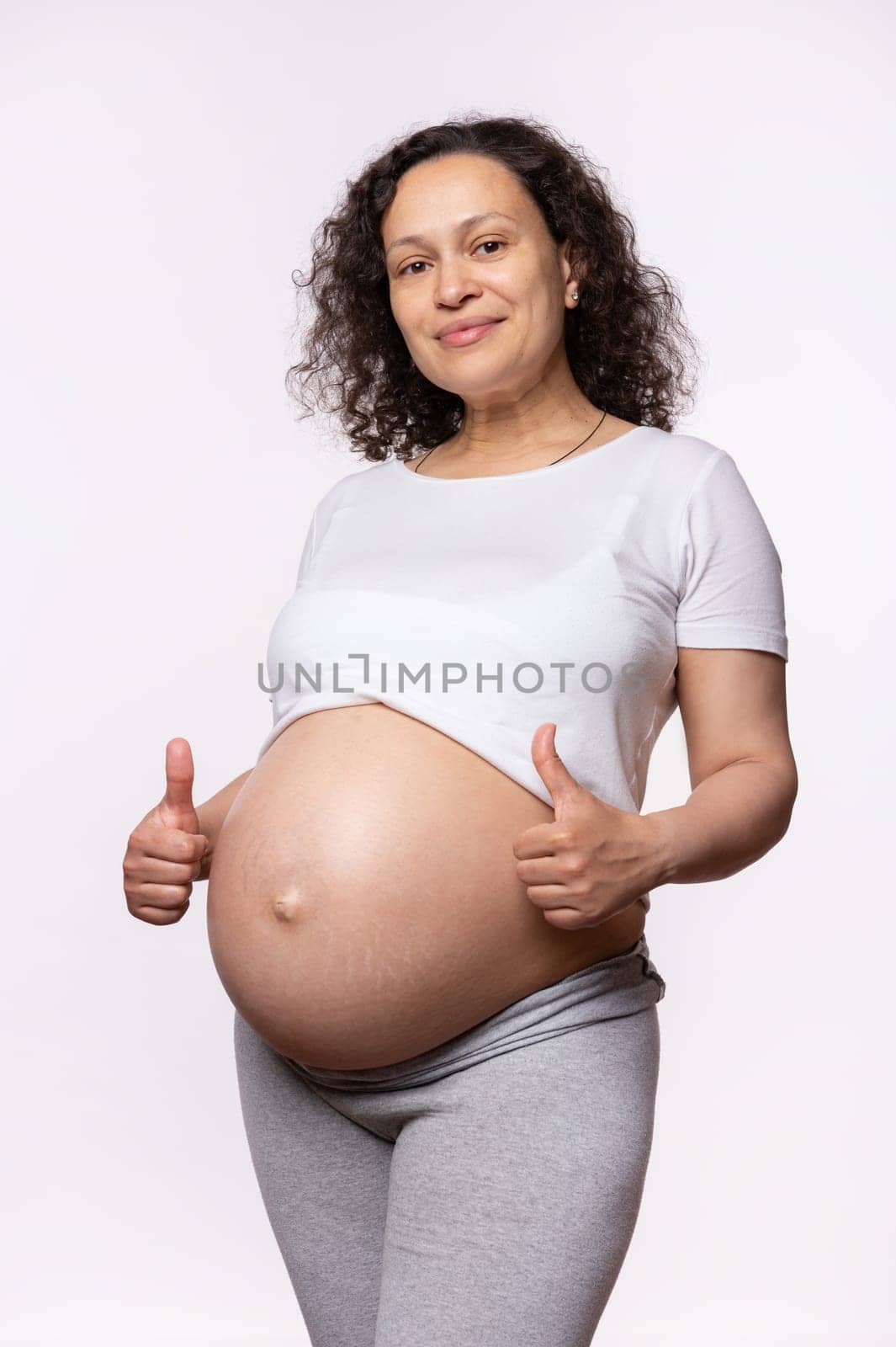 Multi ethnic curly beautiful pregnant woman gestures with thumbs up, expressing happiness and positive emotions, shows approval hand sign, smiles looking at camera, isolated on white studio background