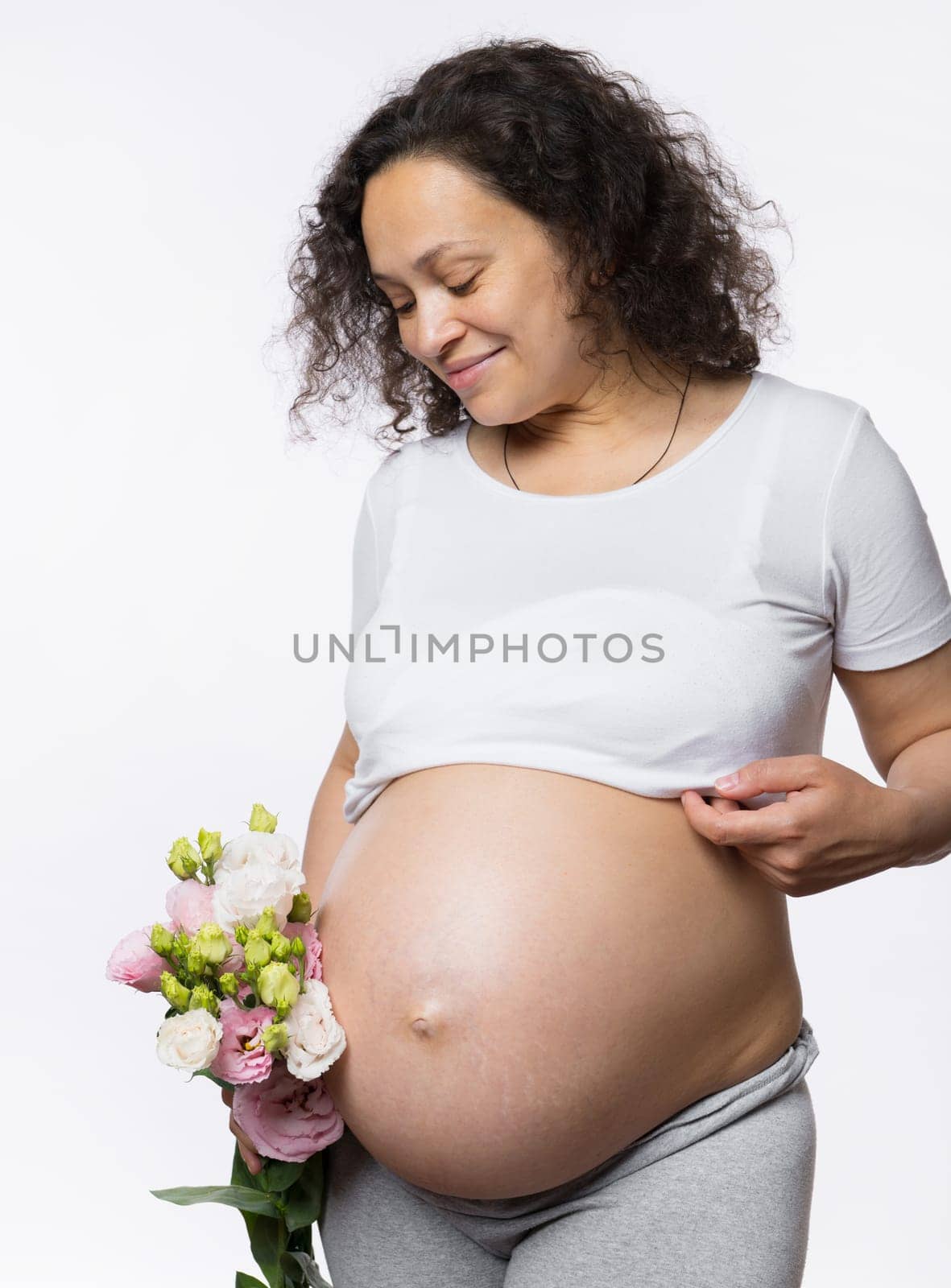 Beautiful multi ethnic happy gravid woman, expectant adult mother holding a bunch of spring flowers, smiling looking at her pregnant belly in late pregnancy time, isolated over white studio background