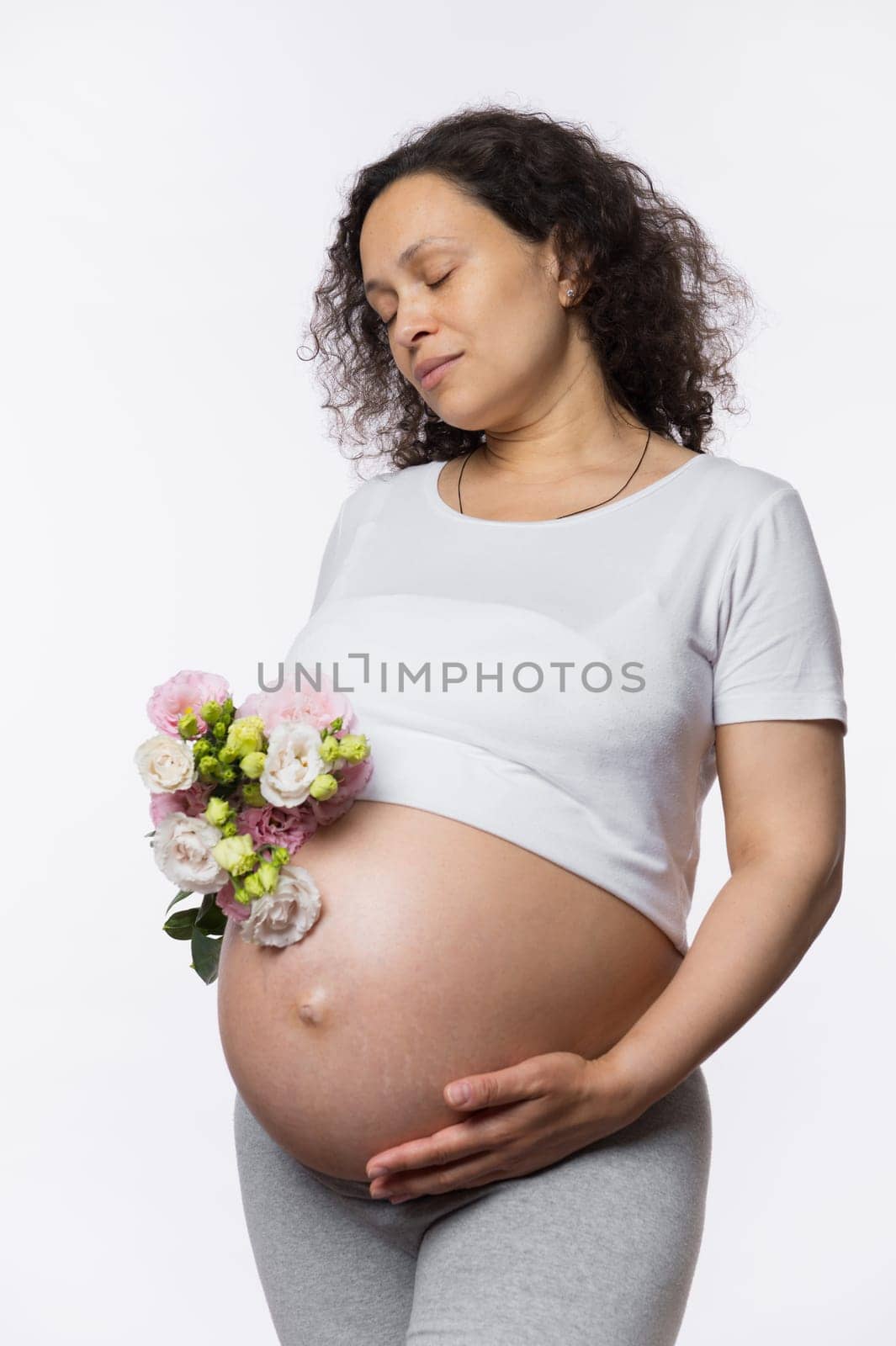 Studio portrait serene pretty pregnant woman with her eyes closed, holding bouquet of flowers in hand near her belly, gently stroking her tummy, isolated on white background. Happy pregnancy concept