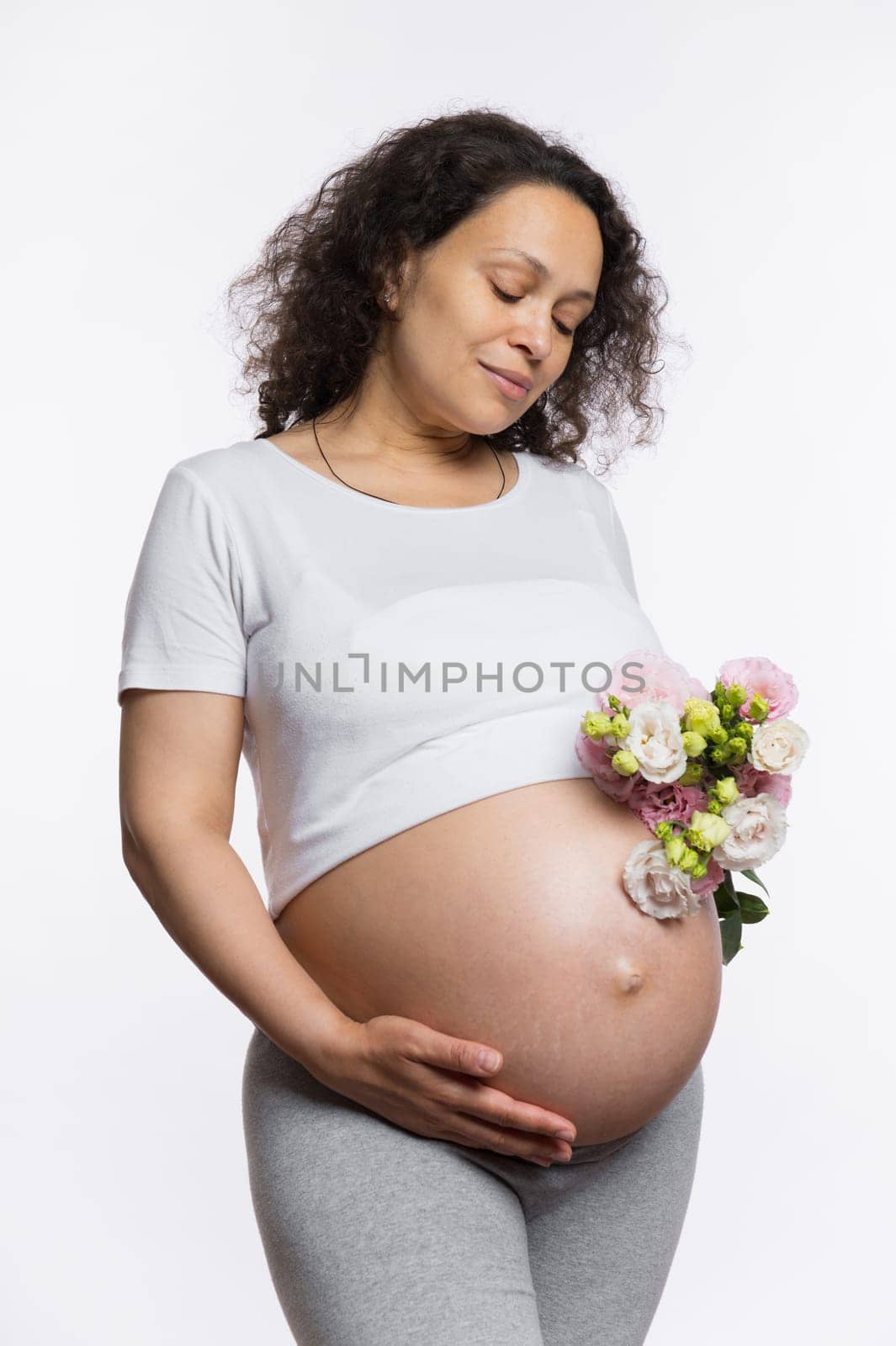 Pregnancy. Motherhood. Mother's Day concept. Beautiful pregnant woman with bunch of flowers, holding hand on her naked belly abdomen, isolated white background. Expectant mother waiting for baby birth