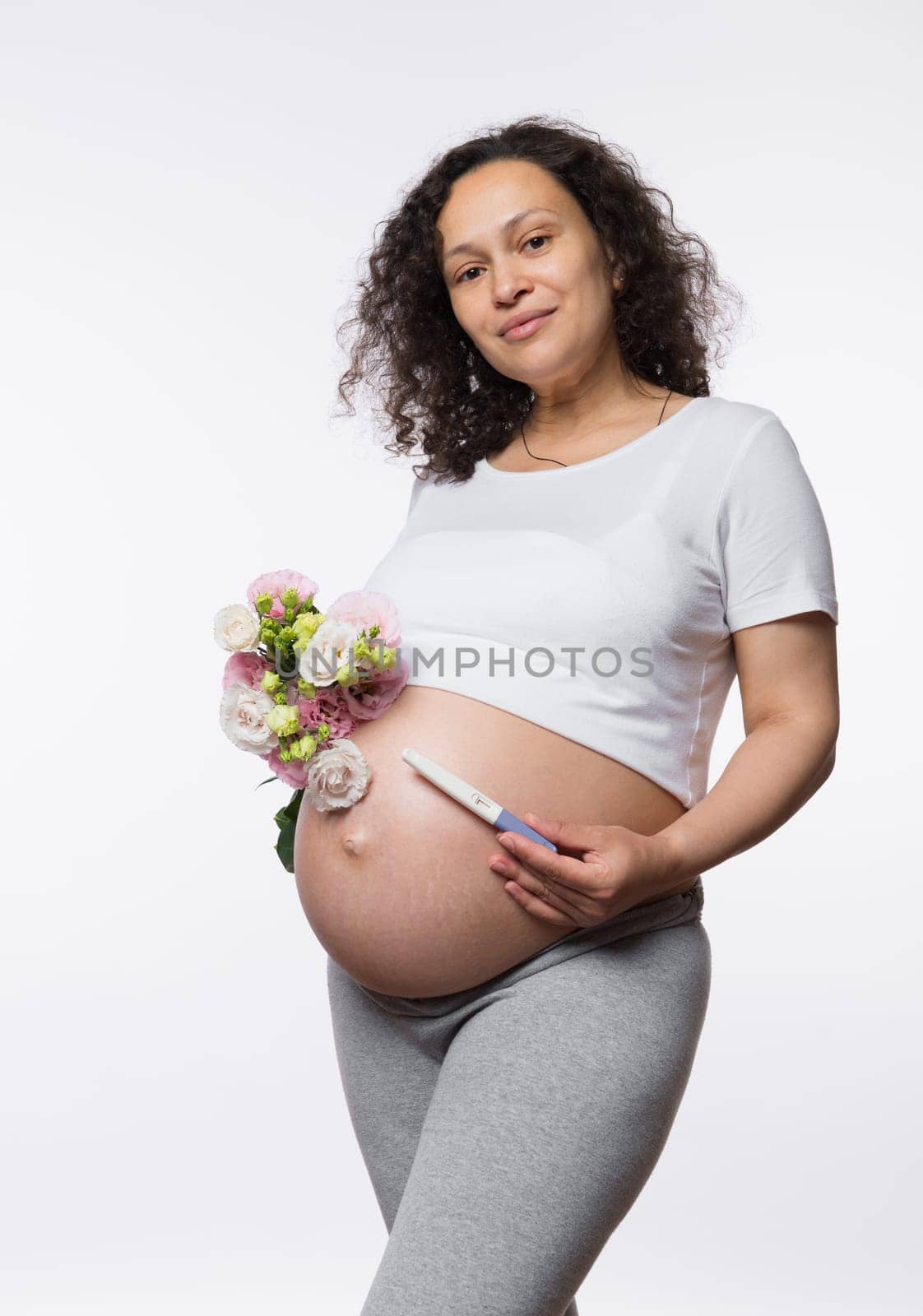 Expecting a baby. Childbearing, childbirth and maternity concept. Beautiful pregnant woman holding pregnancy test and bouquet of flowers, smiling looking at camera, isolated on white studio background
