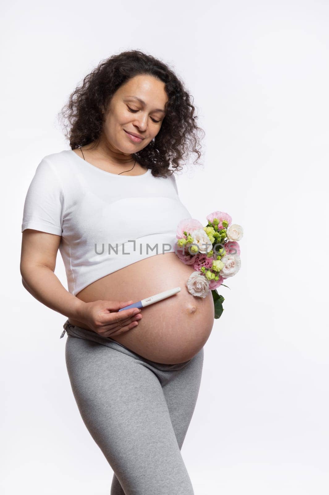 Happy smiling adult gravid woman holding a pregnancy inkjet test kit with positive result and a bouquet of flowers over her big pregnant belly in late pregnancy, isolated over white studio background