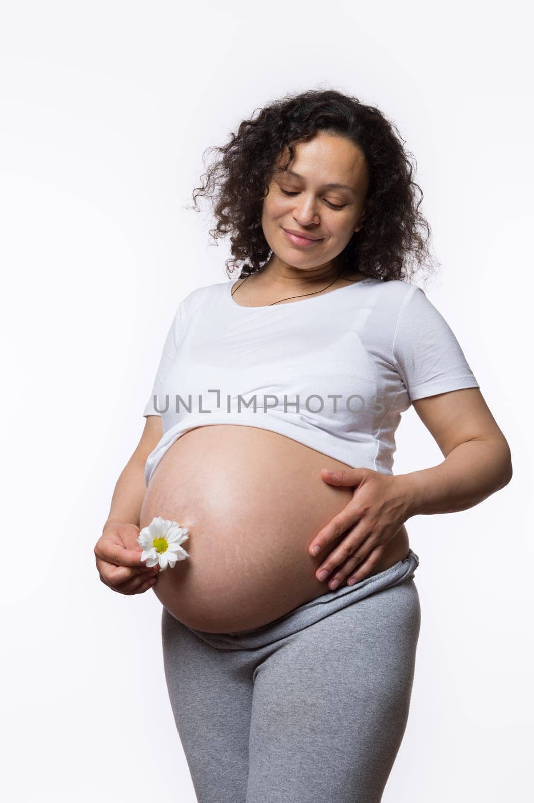 Charming smiling happy Latina gravid mother caressing her big pregnant belly in late pregnancy, holding daisy flower, isolated on white background. Women's health. Obstetrics and gynecology. Maternity