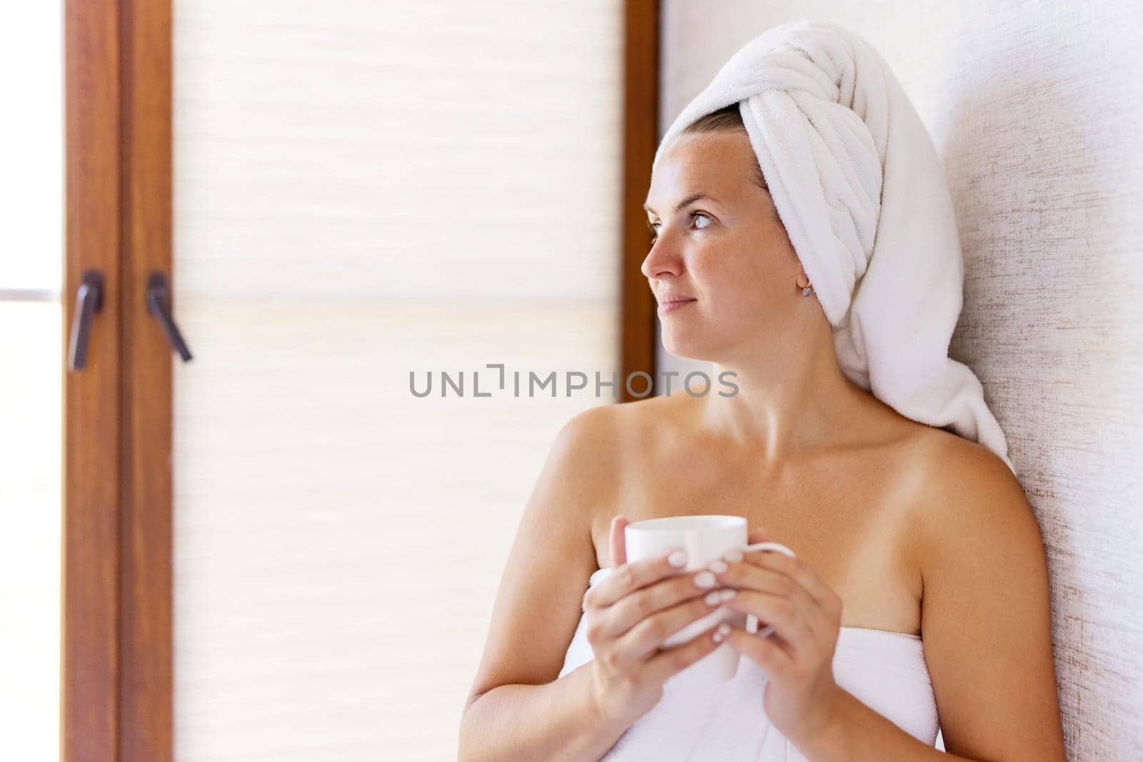 Beautiful young woman with a towel on her head holds a cup of coffee in the morning. Concept of domestic lifestyle.