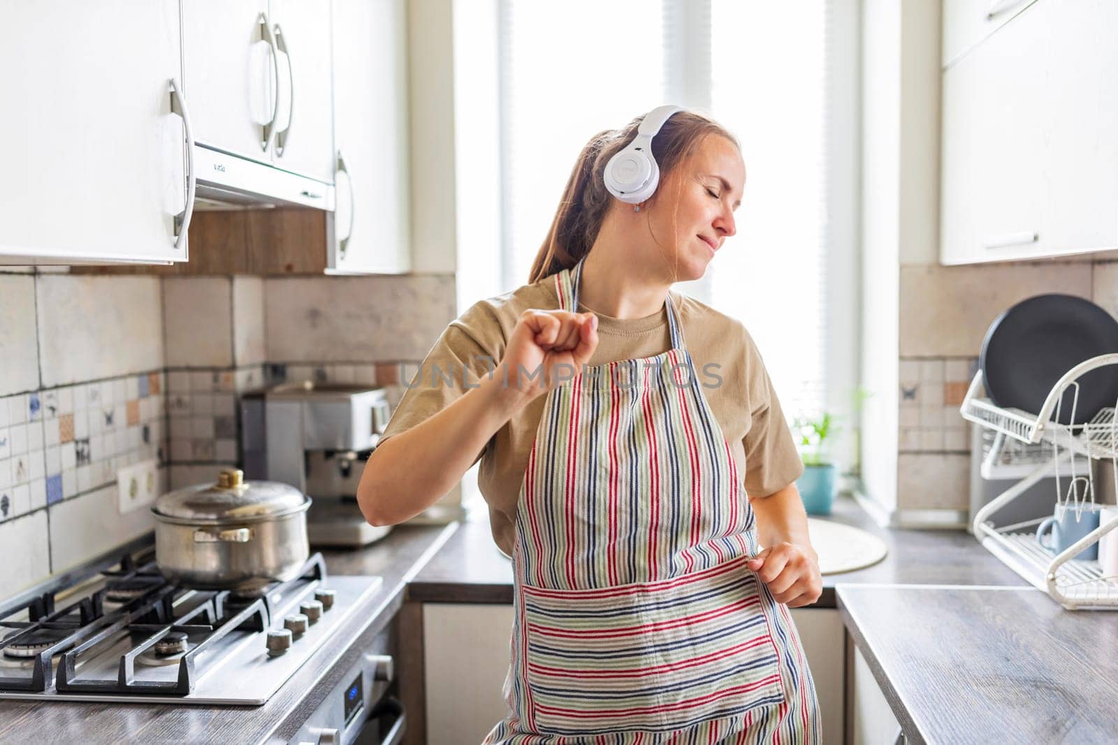 Smiling beautiful woman dancing in kitchen listening music with headphones.