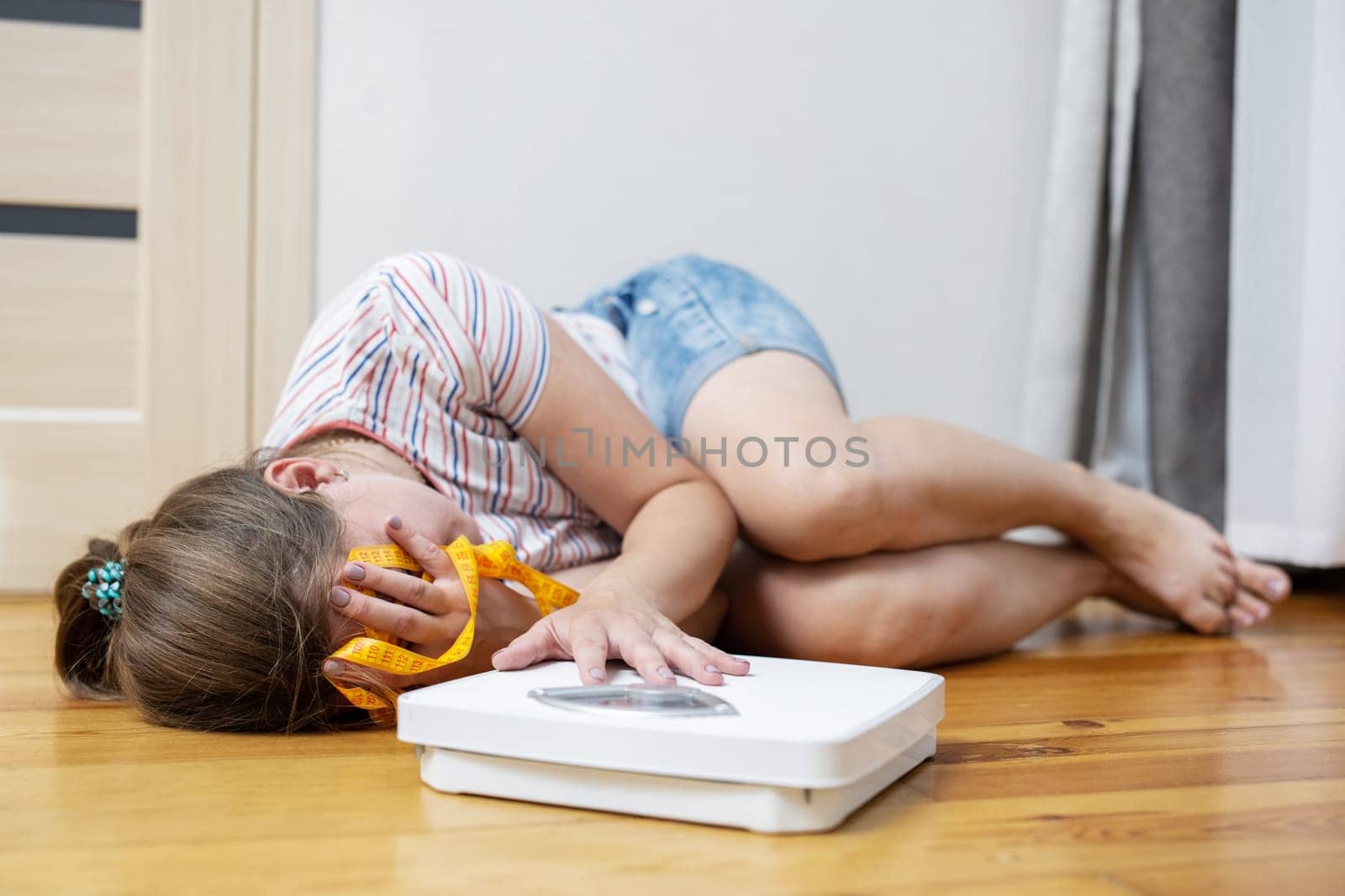 Scales and depressed young woman lying on the floor holding her head and covering her face with her hands.