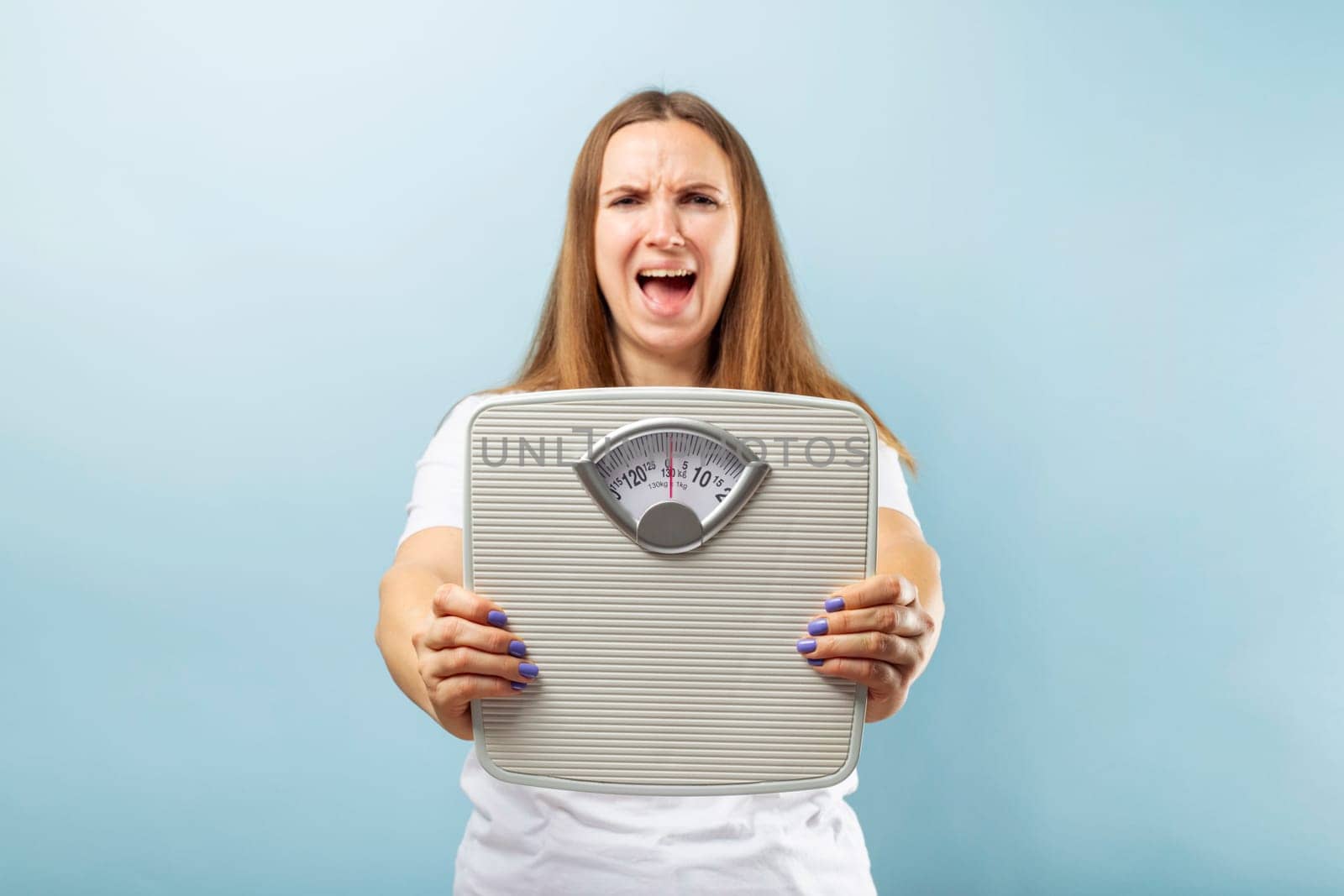 Frustrated young brunette woman in a white t-shirt with weight scale on blue background.