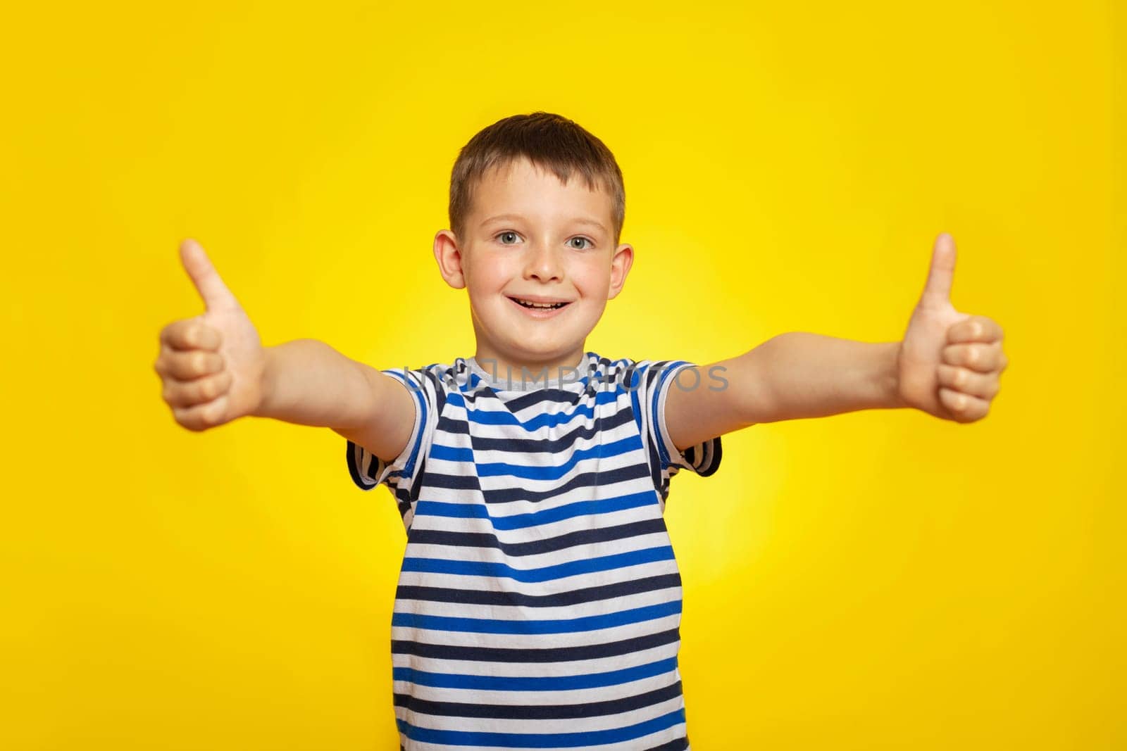 Portrait of happy boy in striped t-shirt showing thumbs up gesture on yellow background.