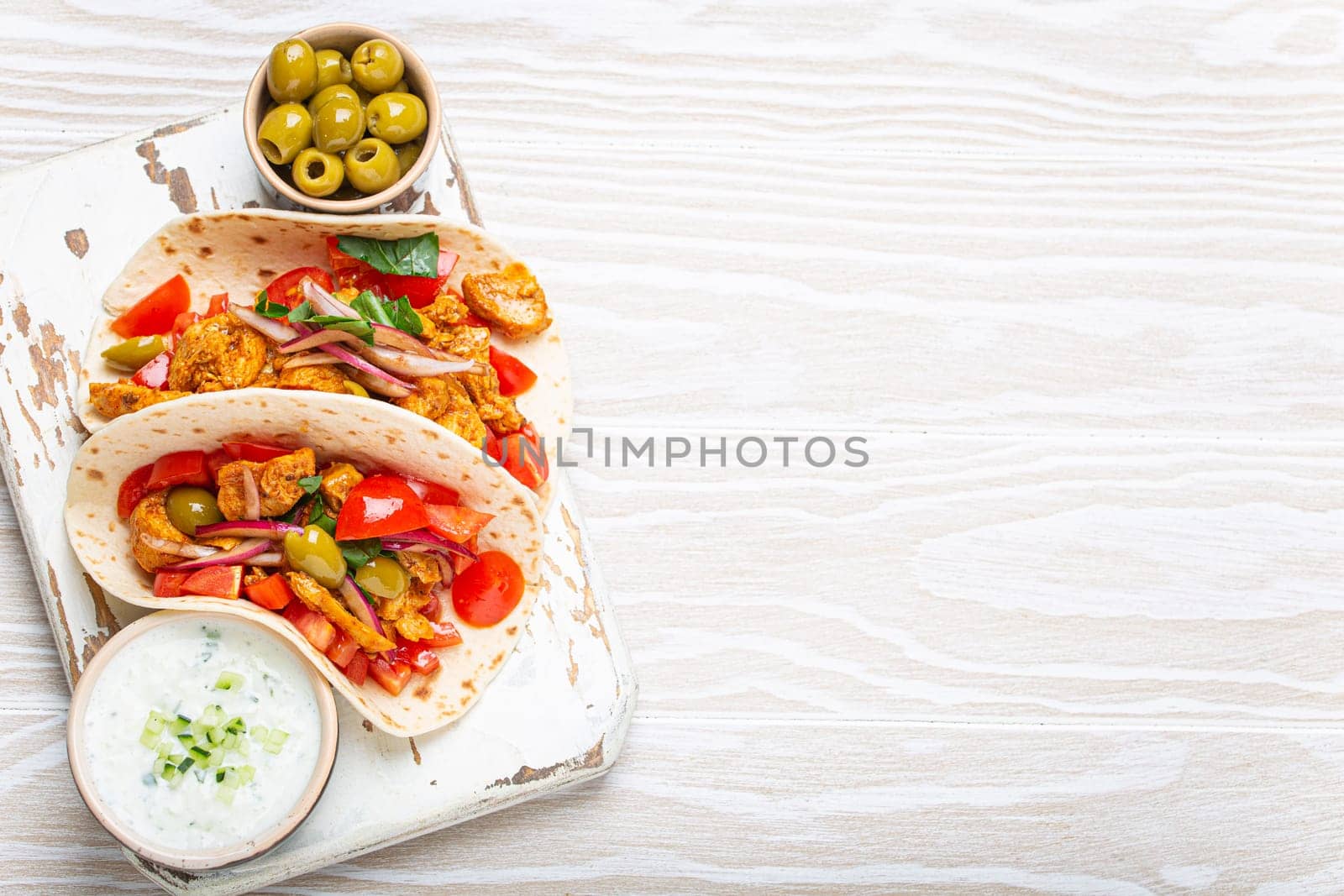 Traditional Greek Dish Gyros: Pita bread Wraps with vegetables, meat, herbs, olives on rustic wooden cutting board with Tzatziki sauce, olive oil top view on white wooden background, space for text.