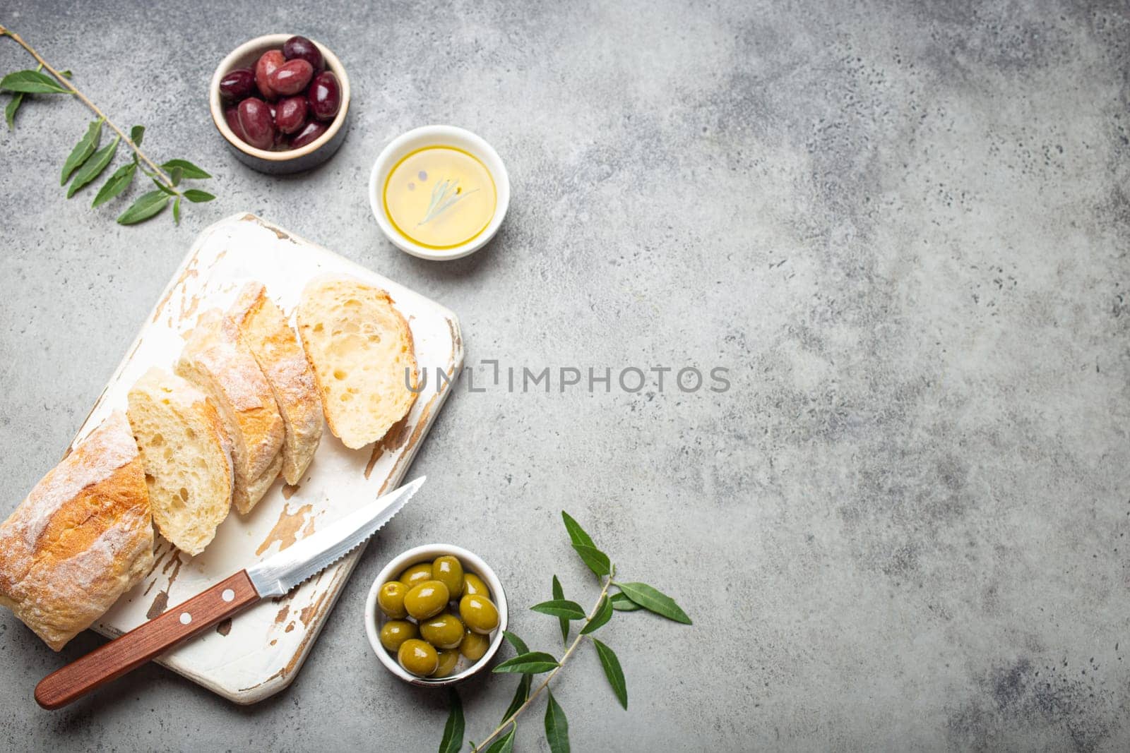 Sliced fresh ciabatta on cutting kitchen board, green and brown olives, olive oil with rosemary, olive tree branches on gray concrete stone rustic background top view copy space by its_al_dente