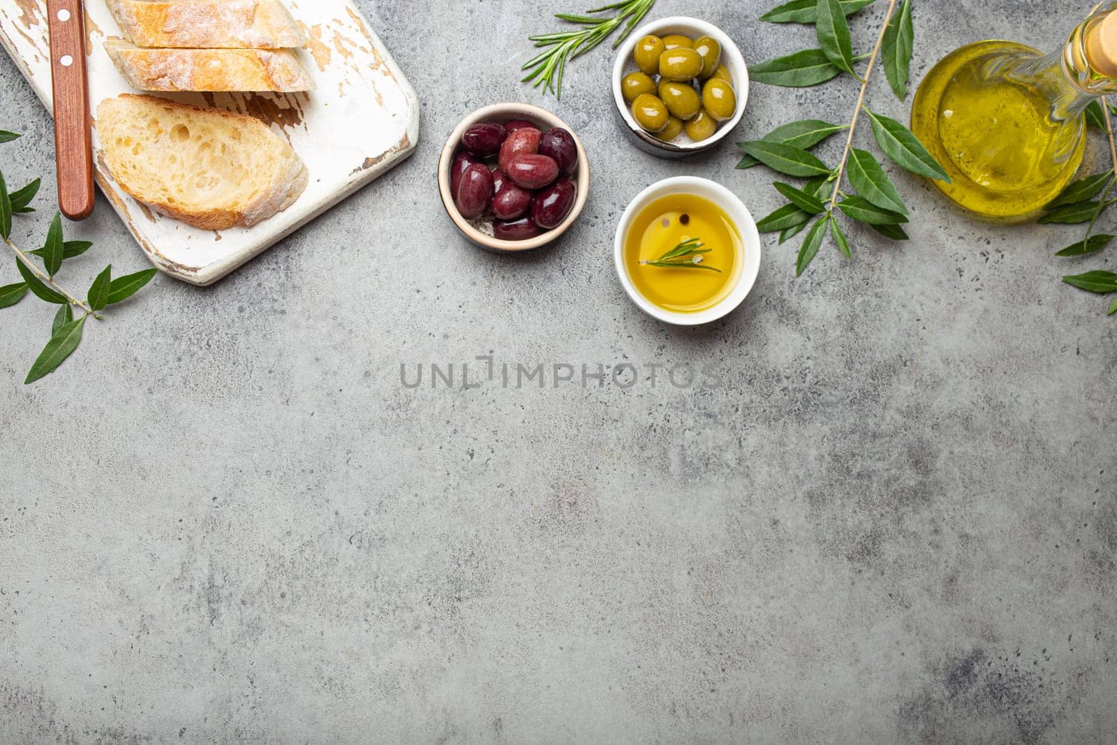 Composition with sliced fresh ciabatta on cutting board, green and brown olives, olive oil with rosemary, olive tree branches on gray concrete stone rustic background top view, copyspace border