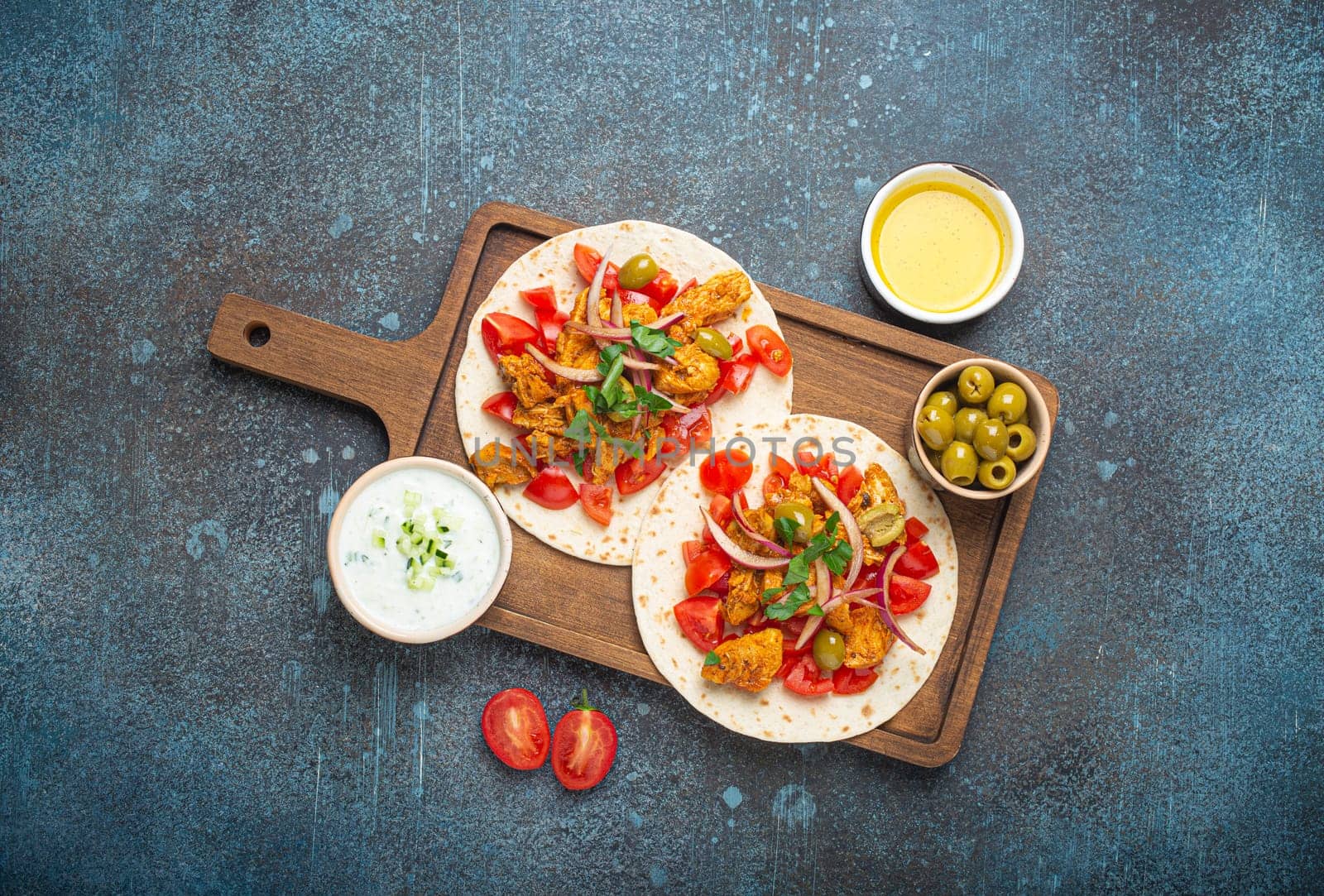 Cooking Traditional Greek Dish Gyros: Pita bread with vegetables, meat, herbs, olives on rustic wooden cutting board with Tzatziki sauce, olive oil top view on dark blue stone background.