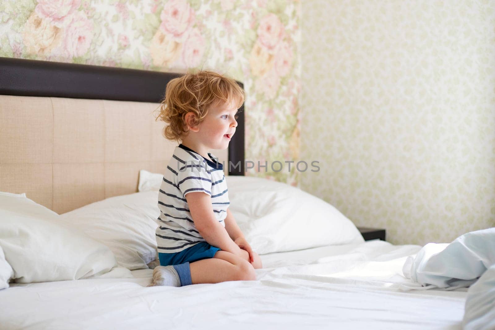 Cute laughing child boy sits in bed. Boy plays at home. Child in a sunny bedroom.