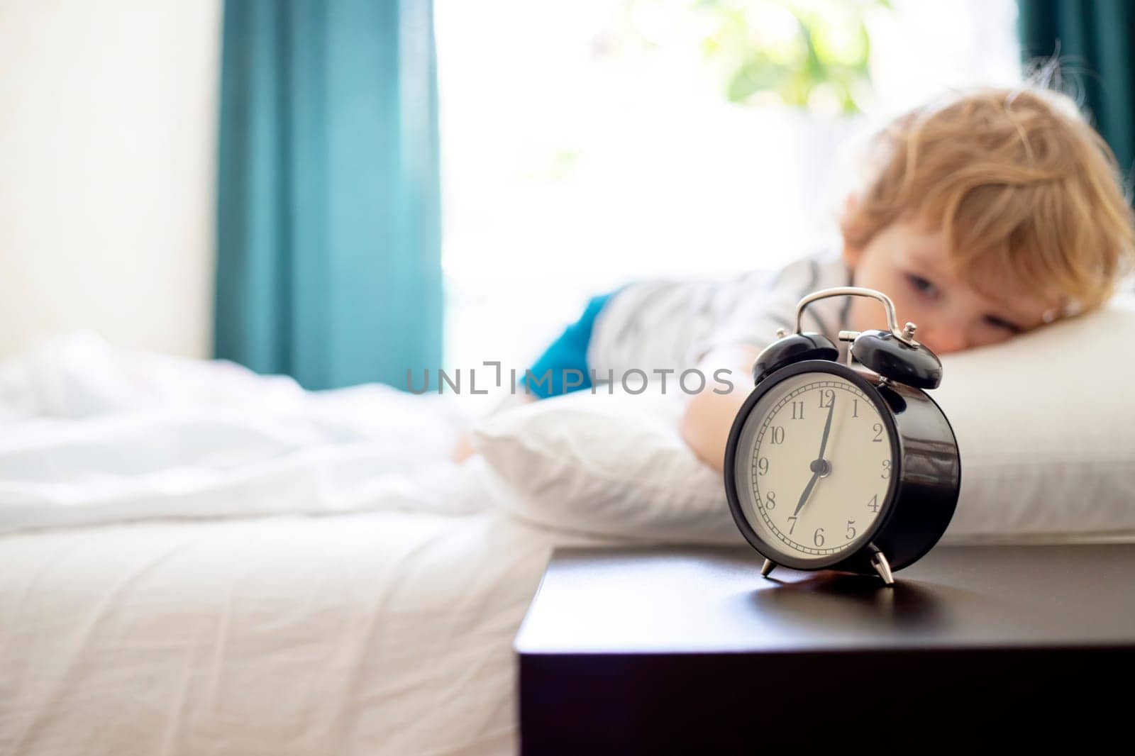 Sleepy little child lies on the bed and reaches for the ringing alarm clock. Sleepy boy on the bed with alarm clock in front.
