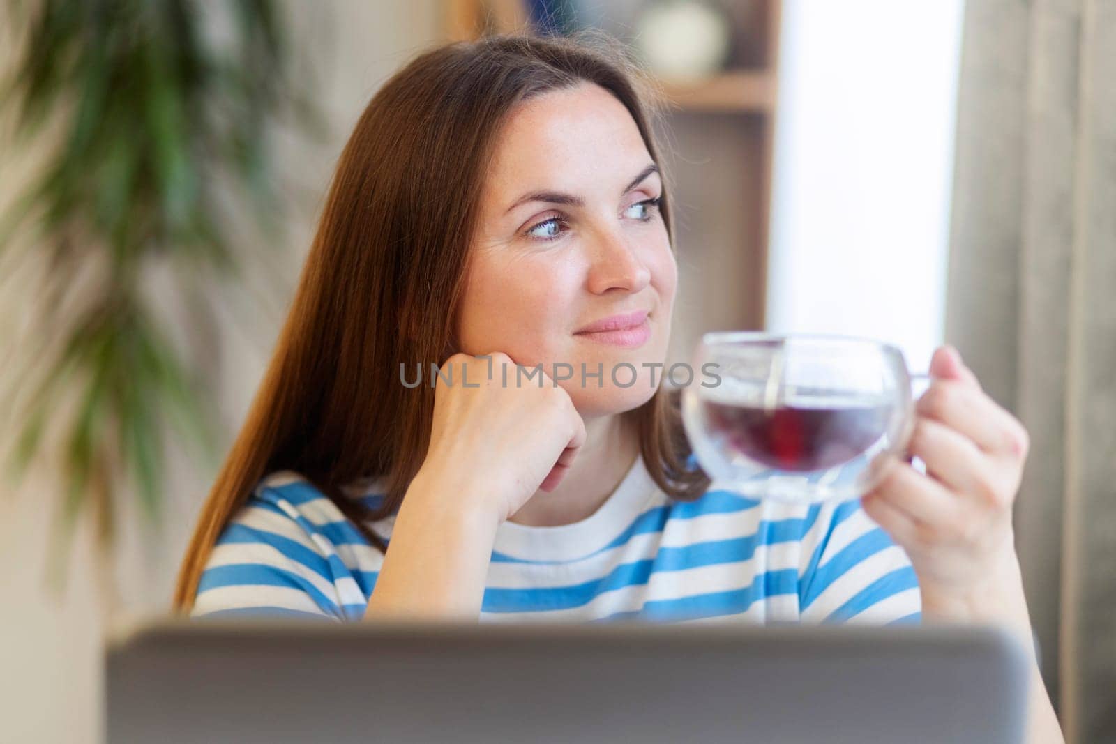 Smiling woman taking a tea break as she sits at a table working on a laptop computer.