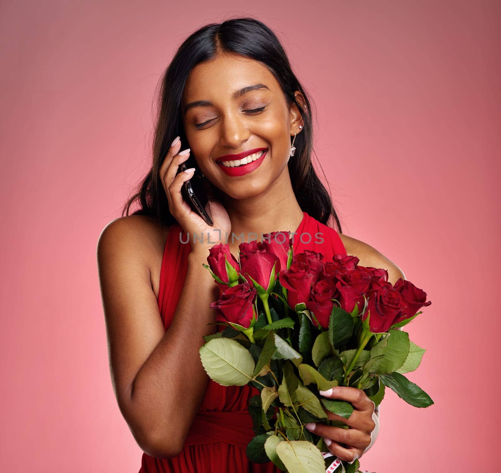 Phone call, talking and a happy woman with flowers on a studio background for valentines day. Laugh, model and face of a young Indian girl with a rose bouquet and smartphone for romance or love.