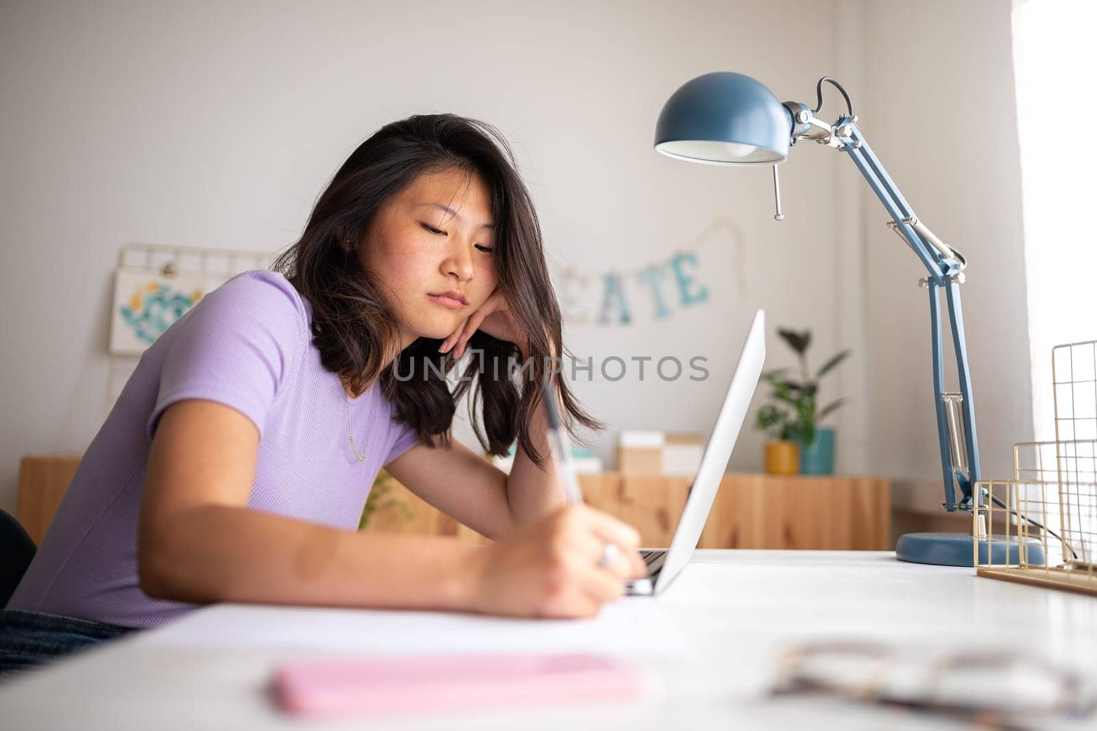 Tired chinese female college student doing homework writing on paper at home. Asian girl studying with laptop. by Hoverstock