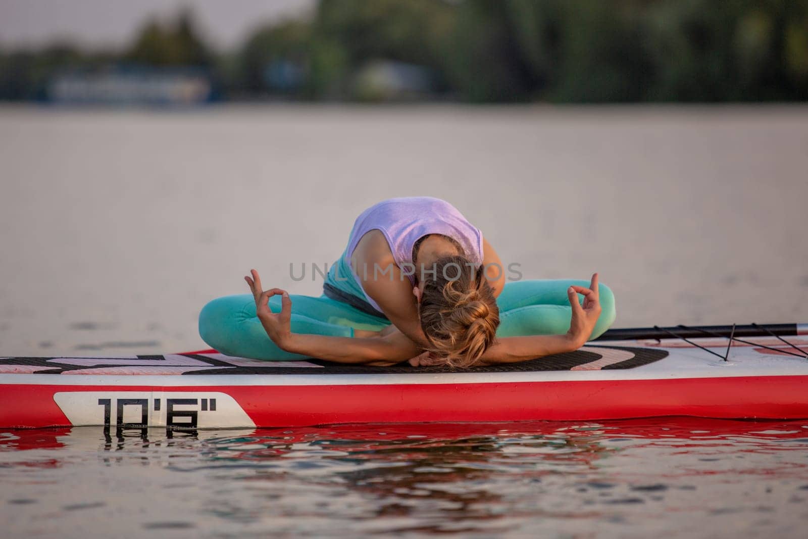 Young woman sitting on paddle board, practicing yoga pose. Doing yoga exercise on sup board, active summer rest. Exercise for flexibility and stretching of muscles.