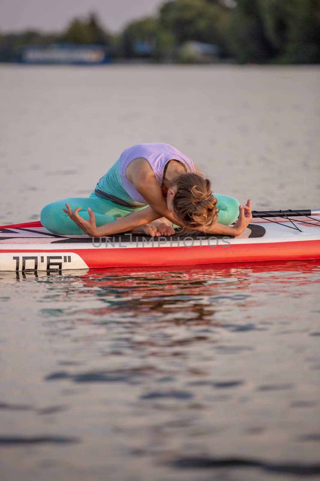 Young woman sitting on paddle board, practicing yoga pose. Doing yoga exercise on sup board, active summer rest. Exercise for flexibility and stretching of muscles.