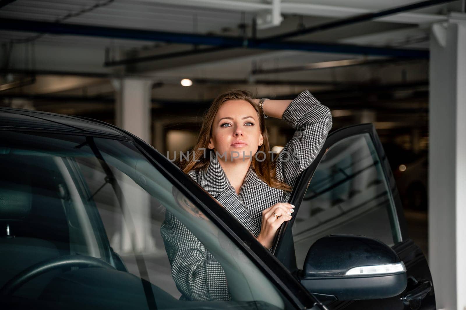 Happy woman car. she stands next to the car in the underground parking. Dressed in a gray coat, holding a glass of coffee in her hands, a black car