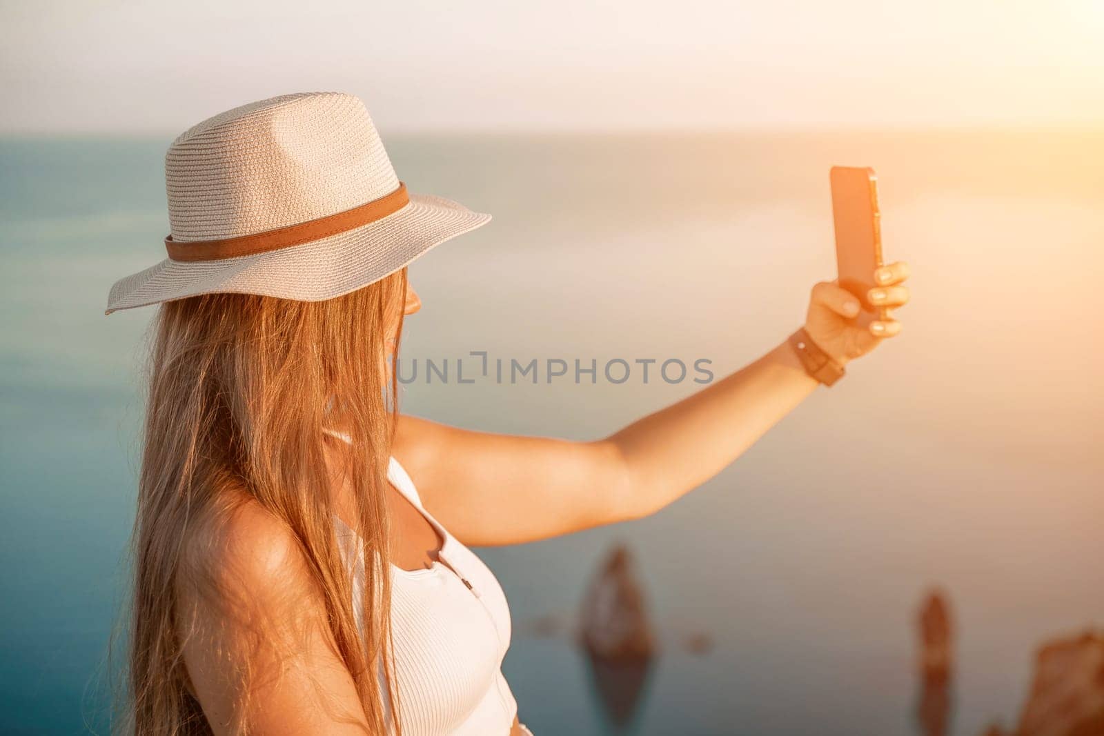 Selfie woman in a hat, white tank top, and shorts captures a selfie shot with her mobile phone against the backdrop of a serene beach and blue sea