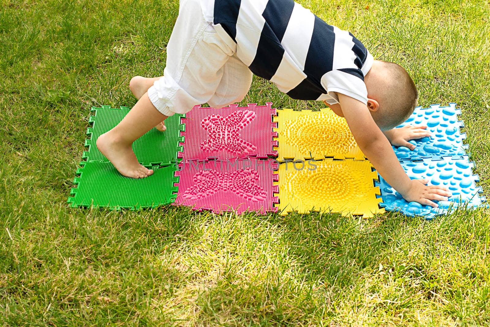 A little boy goes in for sports on massage orthopedic multi-colored rugs. A child in a striped T-shirt crawls merrily in the fresh air in the park, on green grass on massage mats. Stands in a plank position.