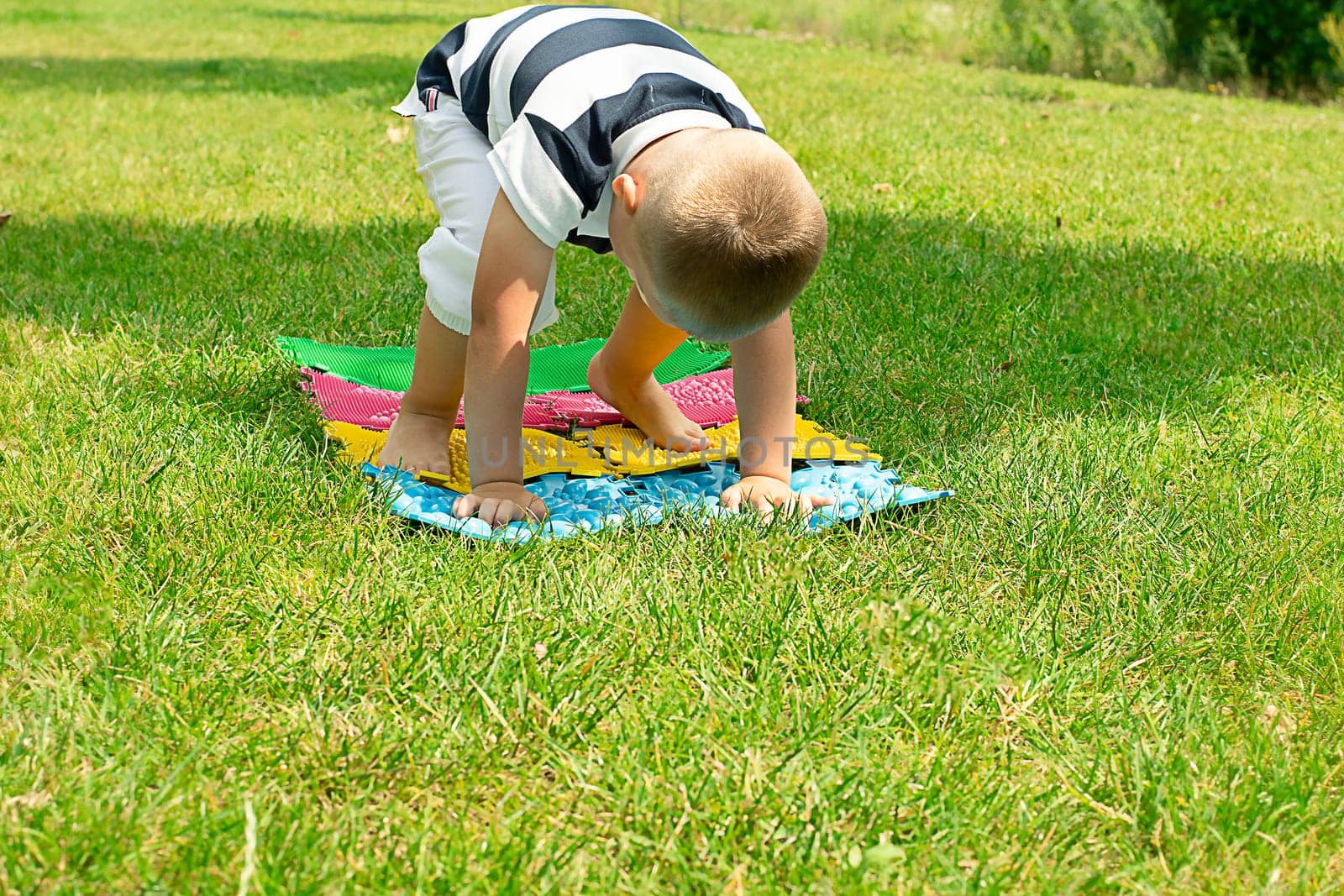 A little boy goes in for sports on massage orthopedic multi-colored rugs. A child in a striped T-shirt crawls merrily in the fresh air in the park, on green grass on massage mats. Stands in a plank position.