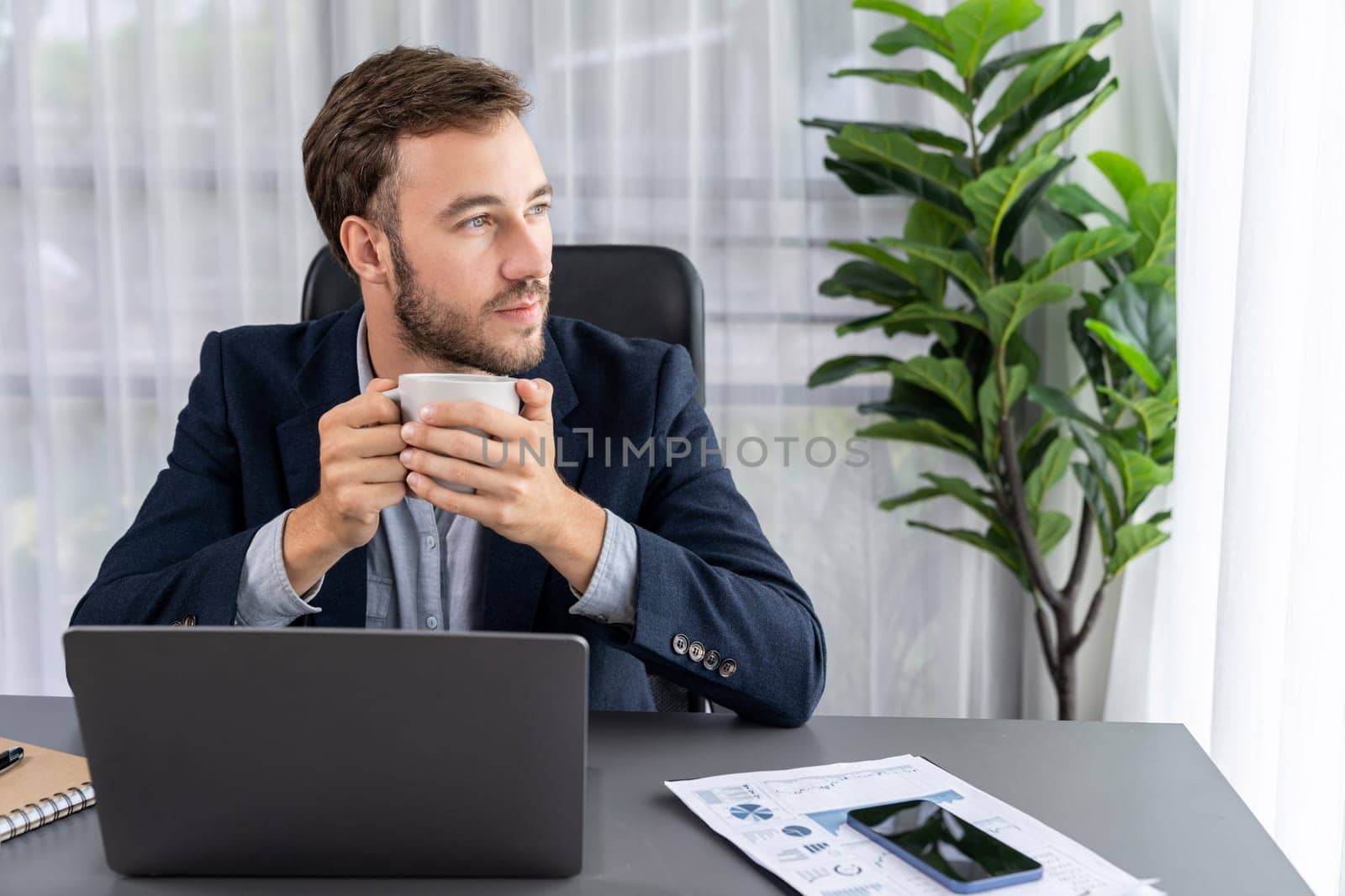 Businessman working on his laptop and drinking coffee at his desk in workspace. Smart executive taking coffee break after researching financial data and marketing plan on his corporate laptop. Entity