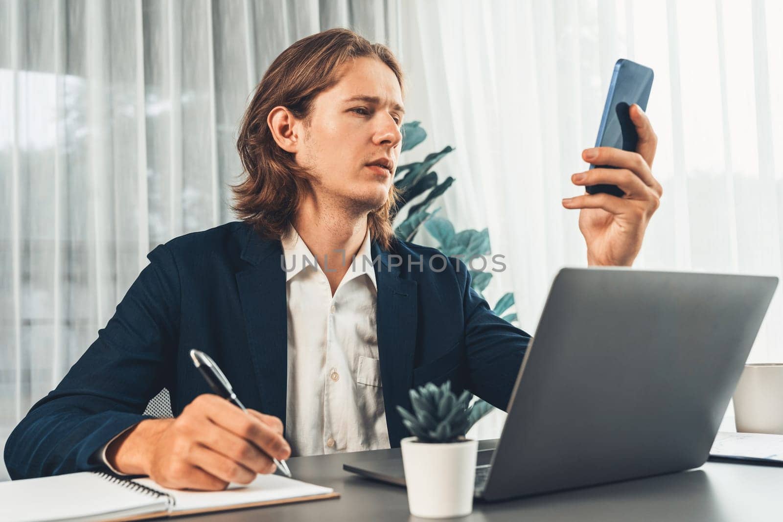 Hardworking and diligent businessman working on his laptop while talking to clients on his mobile phone, showcasing modern multitasking office worker lifestyle. Entity