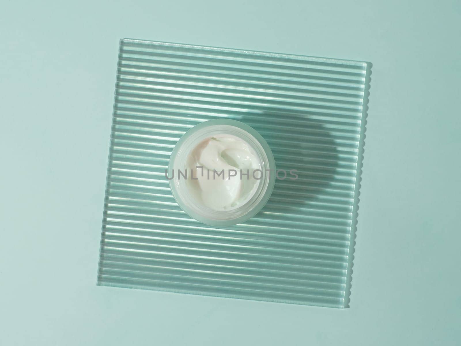 Cosmetic cream or moustirizer on transparent ribbed acrylic plate over blue background. Open round glass jar with aesthetic swirls cream. Top view flatlay. Copy space