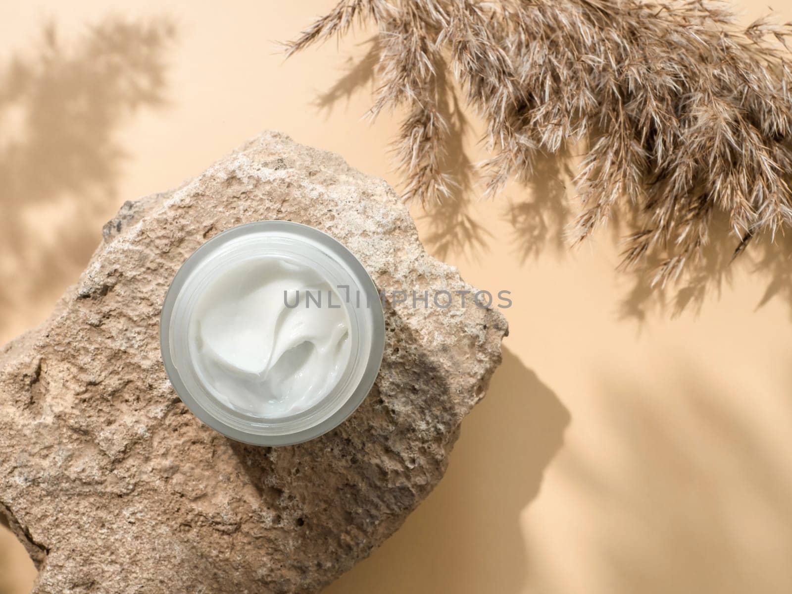 Cosmetic cream or moustirizer on stone, pampas grass on pastel beige background. Beautiful shadows. Open round glass jar with aesthetic swirls face cream. Top view or flat lay. Copy space