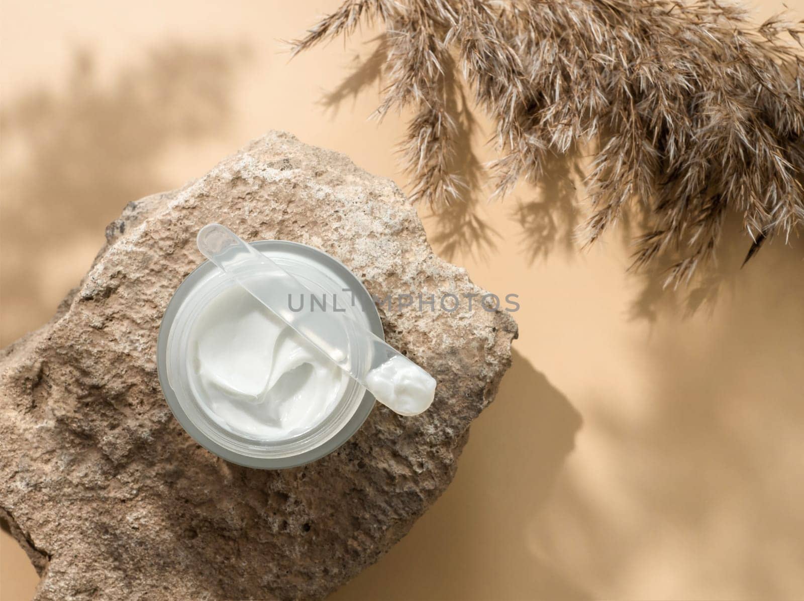 Cosmetic cream or moustirizer on stone, pampas grass on pastel beige background. Beautiful shadows. Open round glass jar with aesthetic swirls face cream and spatula. Flatlay. Copy space