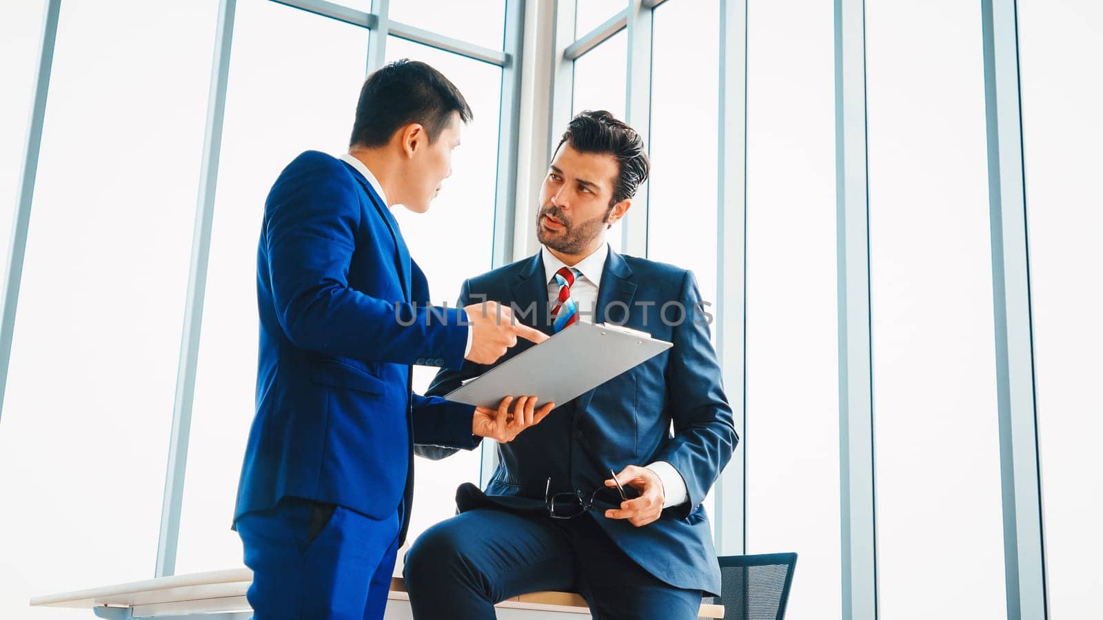 Two business people talk project strategy at office meeting room. Businessman discuss project planning with colleague at modern workplace while having conversation and advice on financial report. Jivy