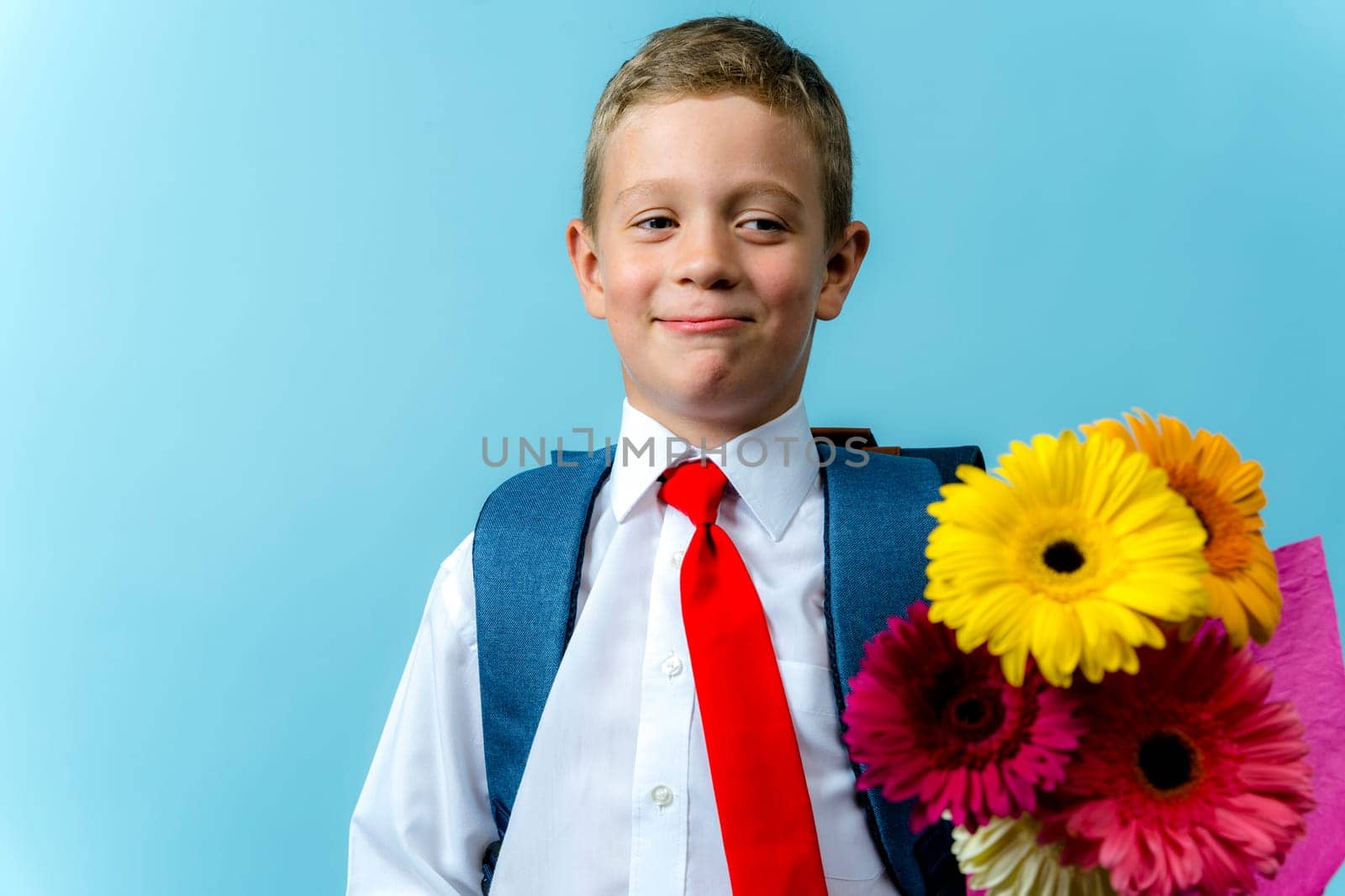 a first grader in a white shirt with a backpack holds a bouquet of flowers in his hands. Cute Caucasian boy goes to school with a bouquet of flowers and a backpack. Schoolboy. September 1, school education. for the first time in the first grade and a bouquet of flowers to the teacher, blue background