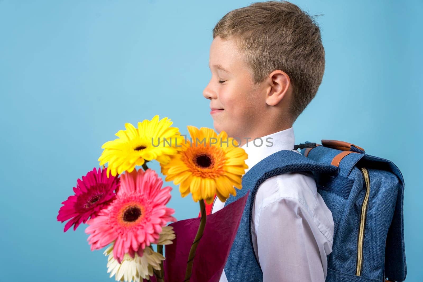 a first grader in a white shirt with a backpack and closed eyes holds a bouquet of flowers in his hands. Cute Caucasian boy goes to school with a bouquet of flowers and a backpack. Side view. Schoolboy. September 1, for the first time in the first class and, blue background