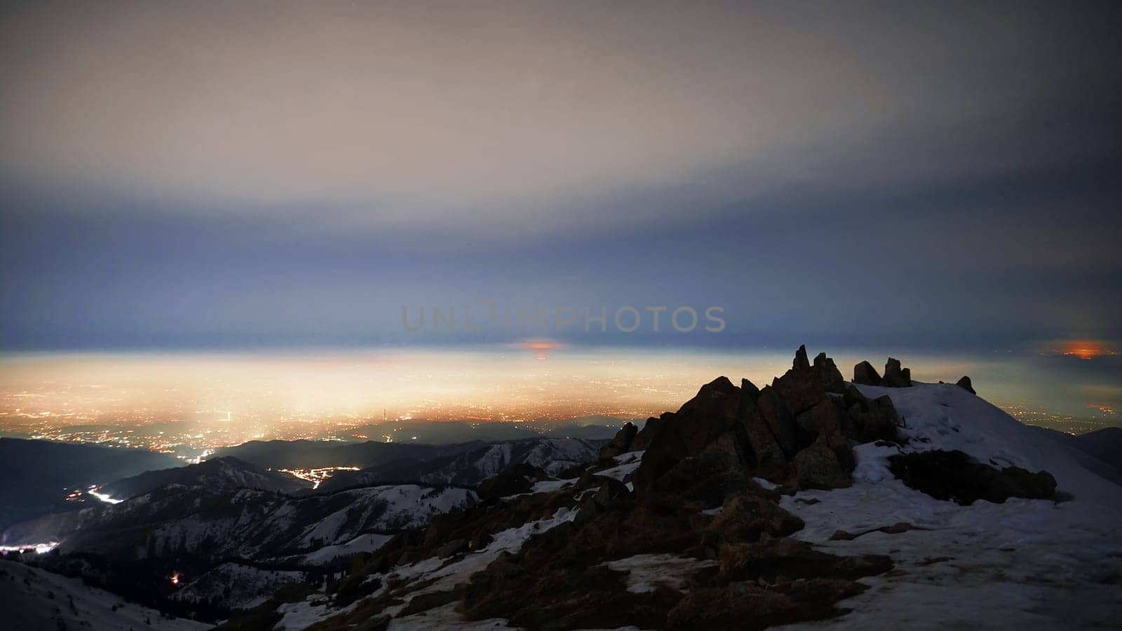 View of the night city from the snowy mountains. The glow from the city rises to the clouds. Bright lights are visible. In places, even the stars are visible. Steep cliffs, big rocks and snow. Almaty