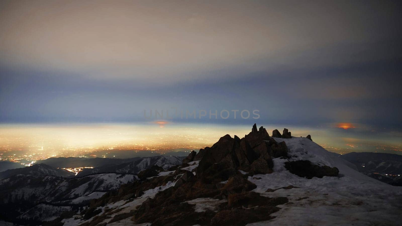 View of the night city from the snowy mountains. The glow from the city rises to the clouds. Bright lights are visible. In places, even the stars are visible. Steep cliffs, big rocks and snow. Almaty