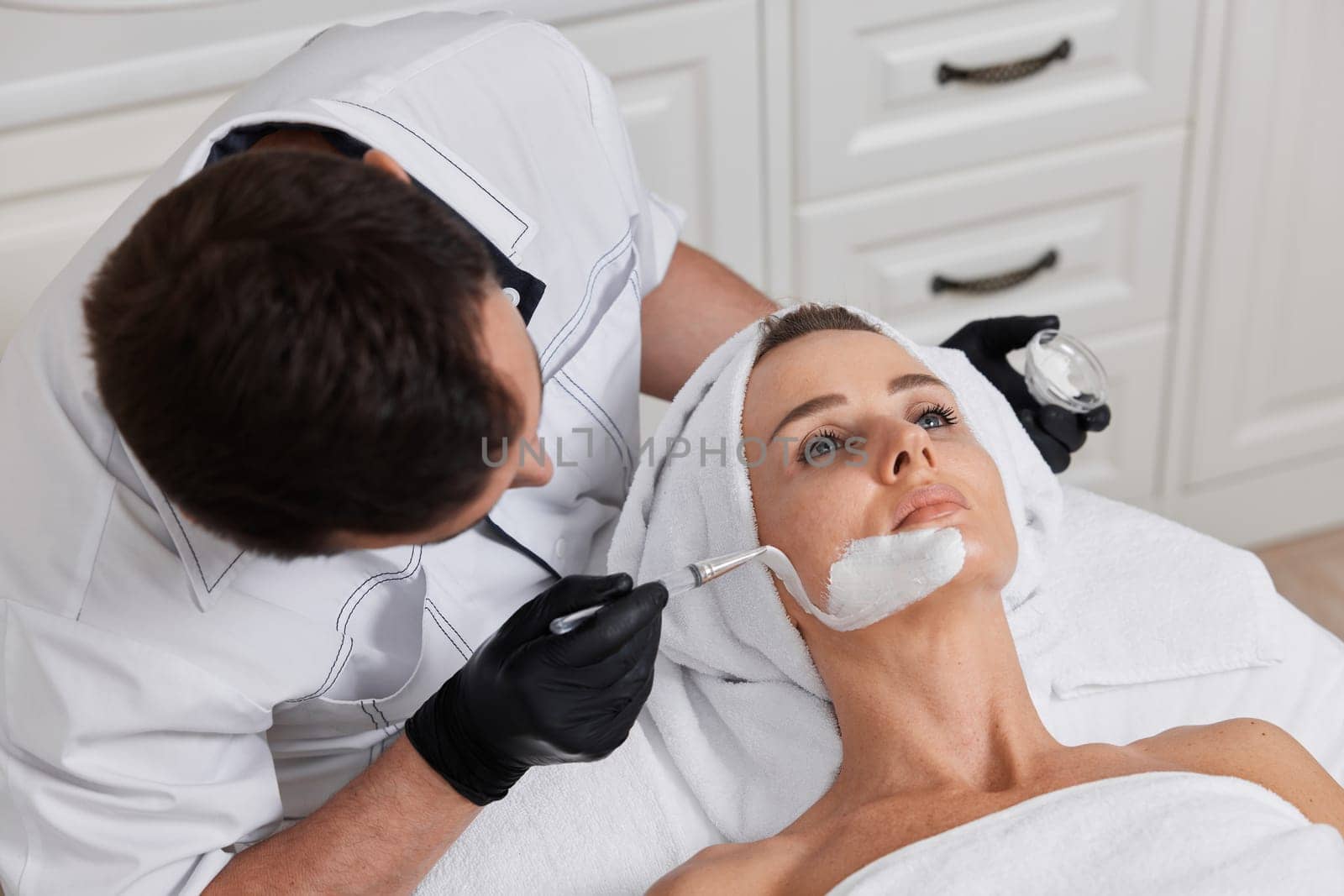 male beautician applies the mask to the face of beautiful woman in the spa salon. facial skin care concept.