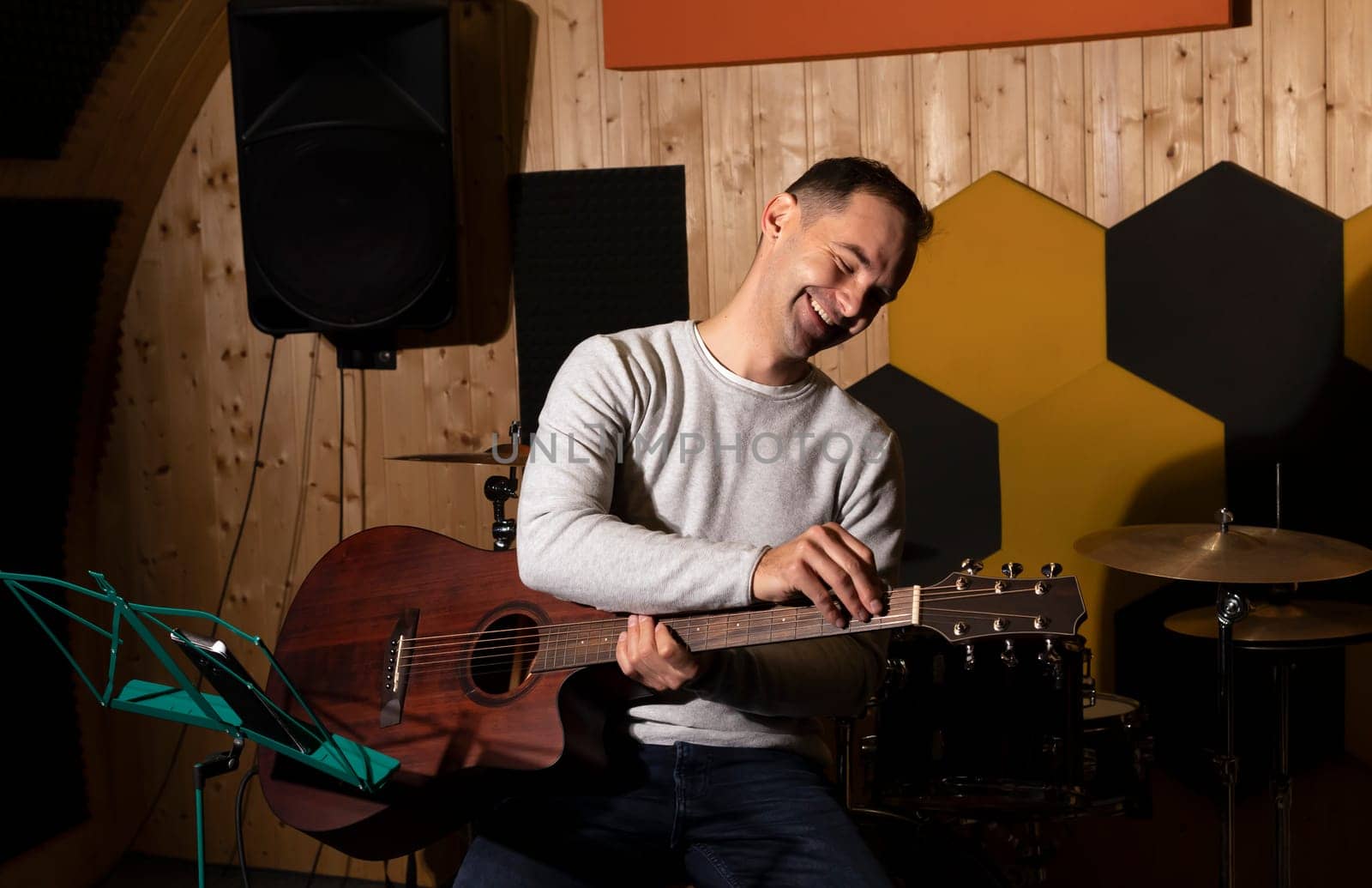 Smiling white man tuning guitar, playing, holding musical instrument in hands, sitting on chair in studio. Darken photo. Male wears casual cloth. Hobby, leisure of creative person, artist.Horizontal
