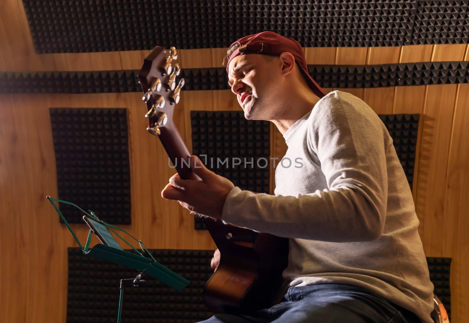 Caucasian man sings song, playing guitar, holding musical instrument in hands, sitting in chair in studio. Darken photo. Male wears casual cloth. Hobby, leisure of creative person, artist.Horizontal