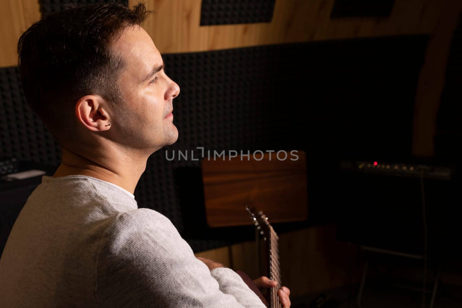 Caucasian man plays guitar, holding musical instrument in hands, sitting in chair in studio. Darken photo. Male wears casual cloth. Hobby, leisure of creative person, artist. Copy space. Horizontal