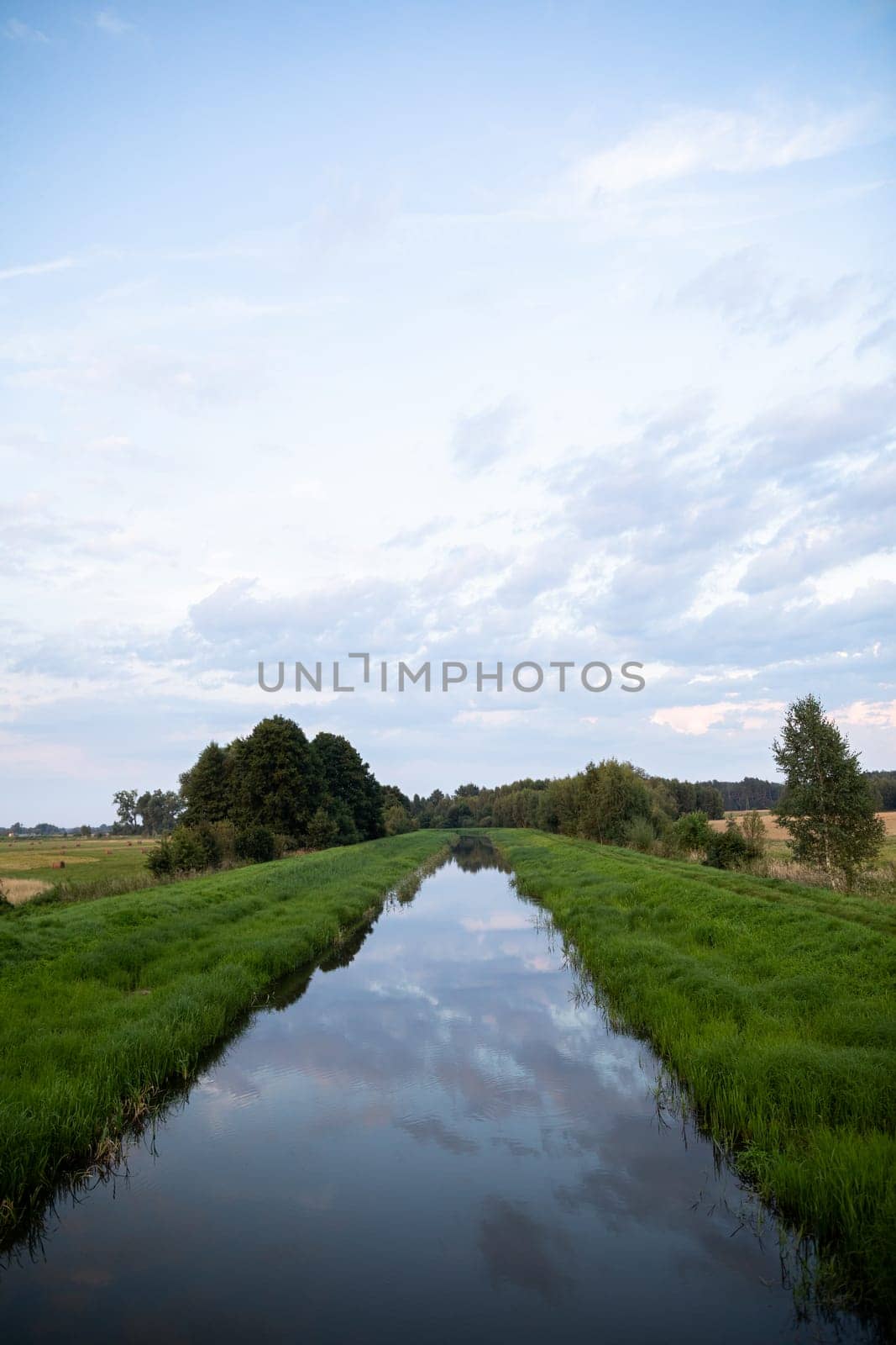The river with a very slow current flows straight amidst green grasses and deciduous trees. The overcast sky is reflected in the calm river.