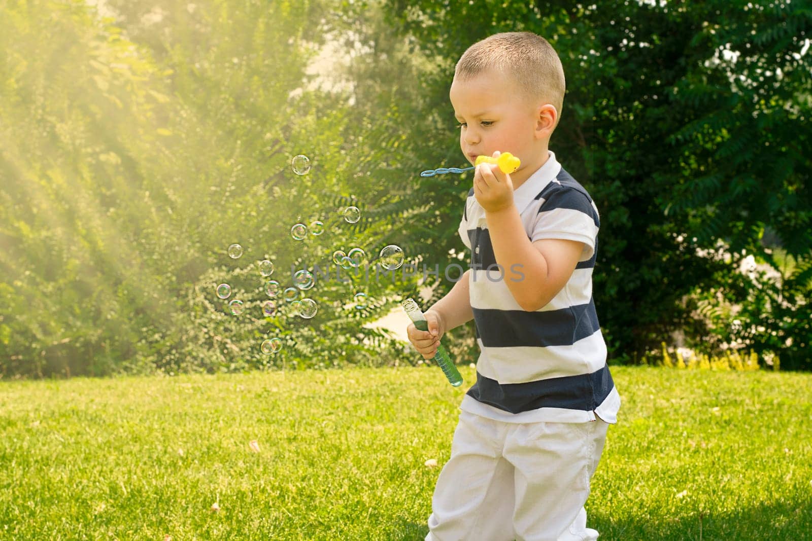 A handsome caucasoid boy 4 years old, in a striped T-shirt, plays outdoors in a green park in summer and blows soap bubbles in the sun. Childhood concept.