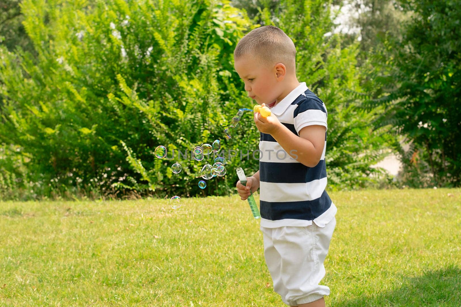A handsome caucasoid boy 4 years old, in a striped T-shirt, plays outdoors in a green park in summer and blows soap bubbles in the sun. Childhood concept.