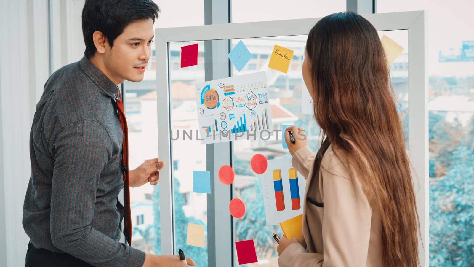 Business people work on project planning board in office and having conversation with coworker friend to analyze project development . They use sticky notes posted on glass to make it organized. Jivy