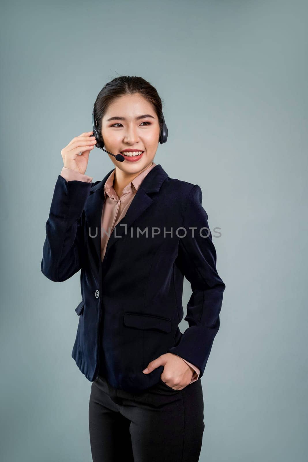 Attractive asian female call center operator with happy smile face advertises job opportunity on empty space, wearing a formal suit and headset on customizable isolated background. Enthusiastic