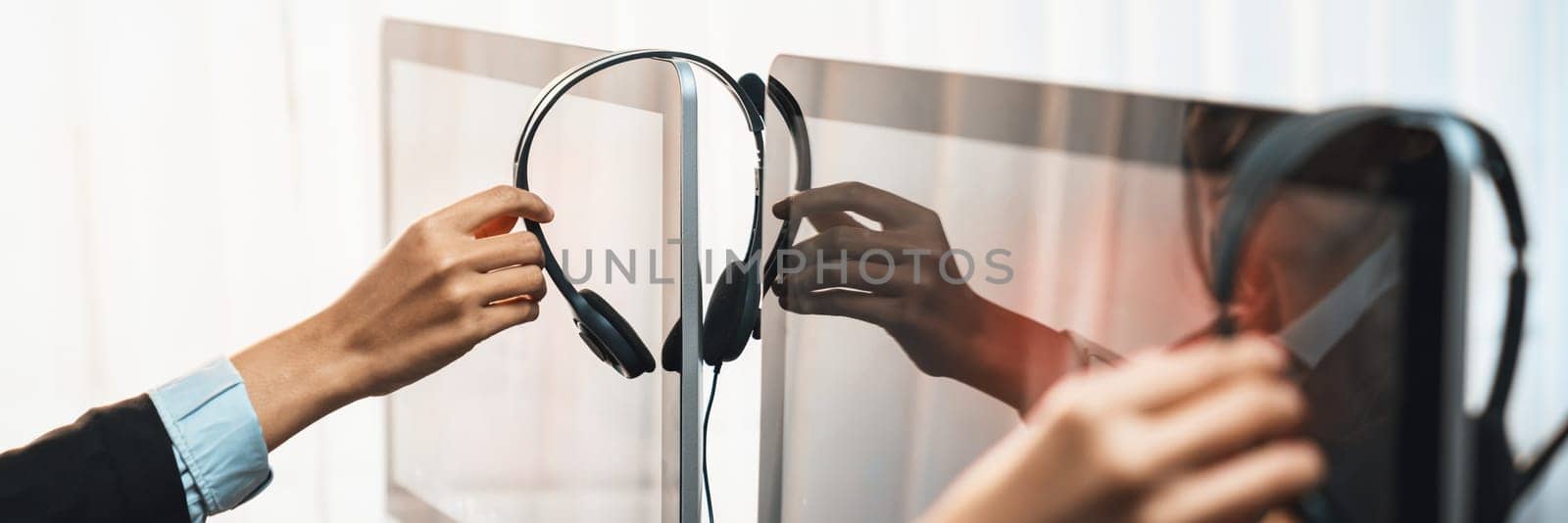 Panorama focus hand holding headset on call center workspace desk with blur background of operator team or telesales representative engaging in providing client with customer support service. Prodigy